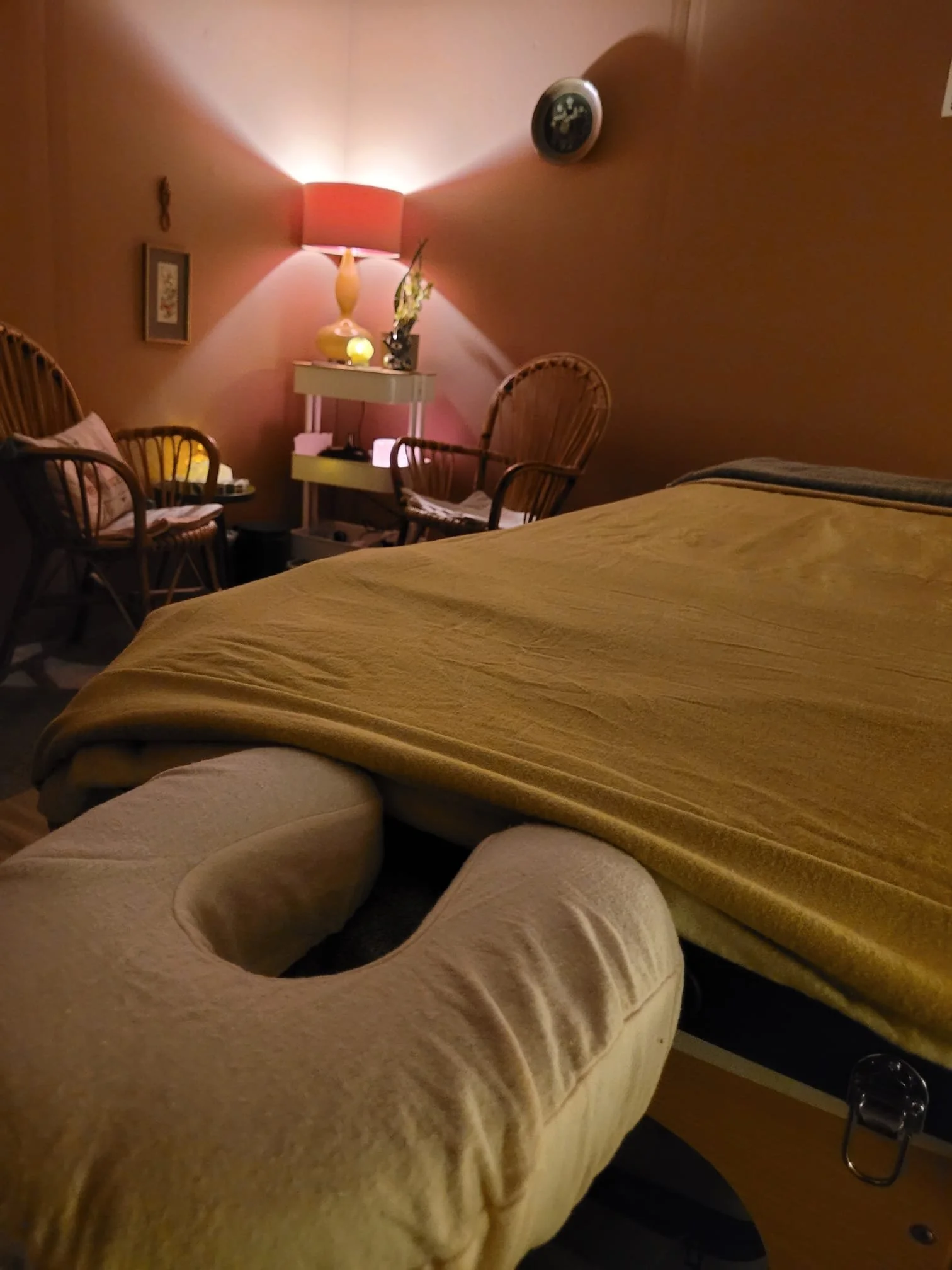 A cozy bedroom with a bed covered in a yellow blanket, a beige armchair, and a sitting area with wicker chairs, a small table with a lamp, and wall decorations. A lamp on the wall illuminates the room with warm light.