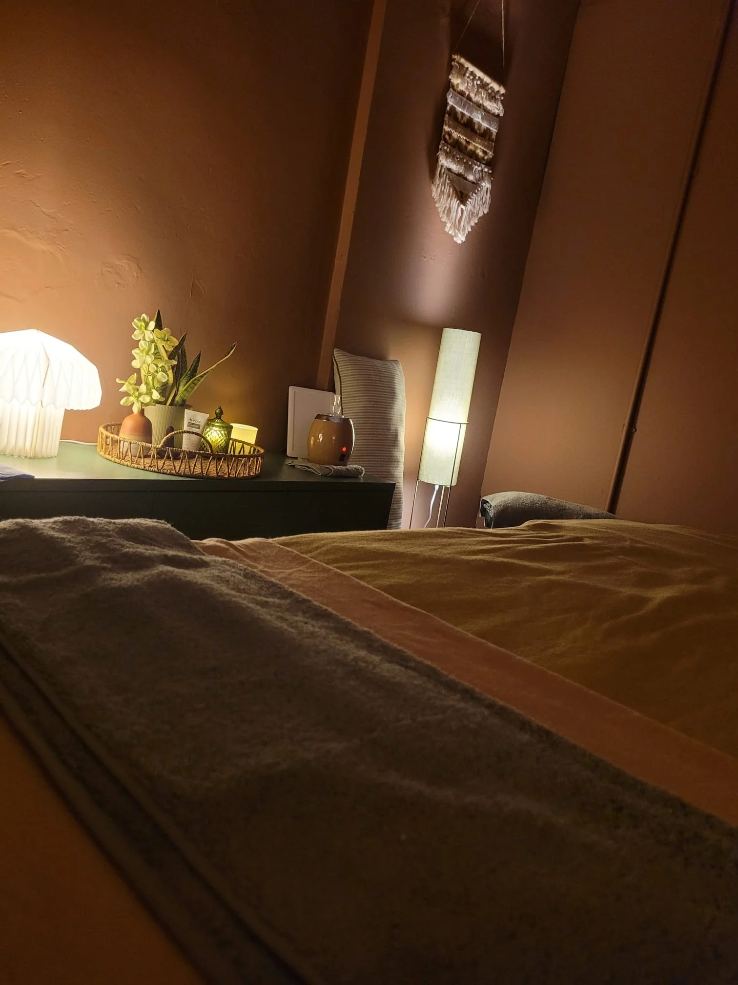 A cozy bedroom with warm lighting, a bed with brown bedding, a nightstand with a lamp, a vase with flowers, a diffuser, and a decorative wall hanging, with a brown wall and window.