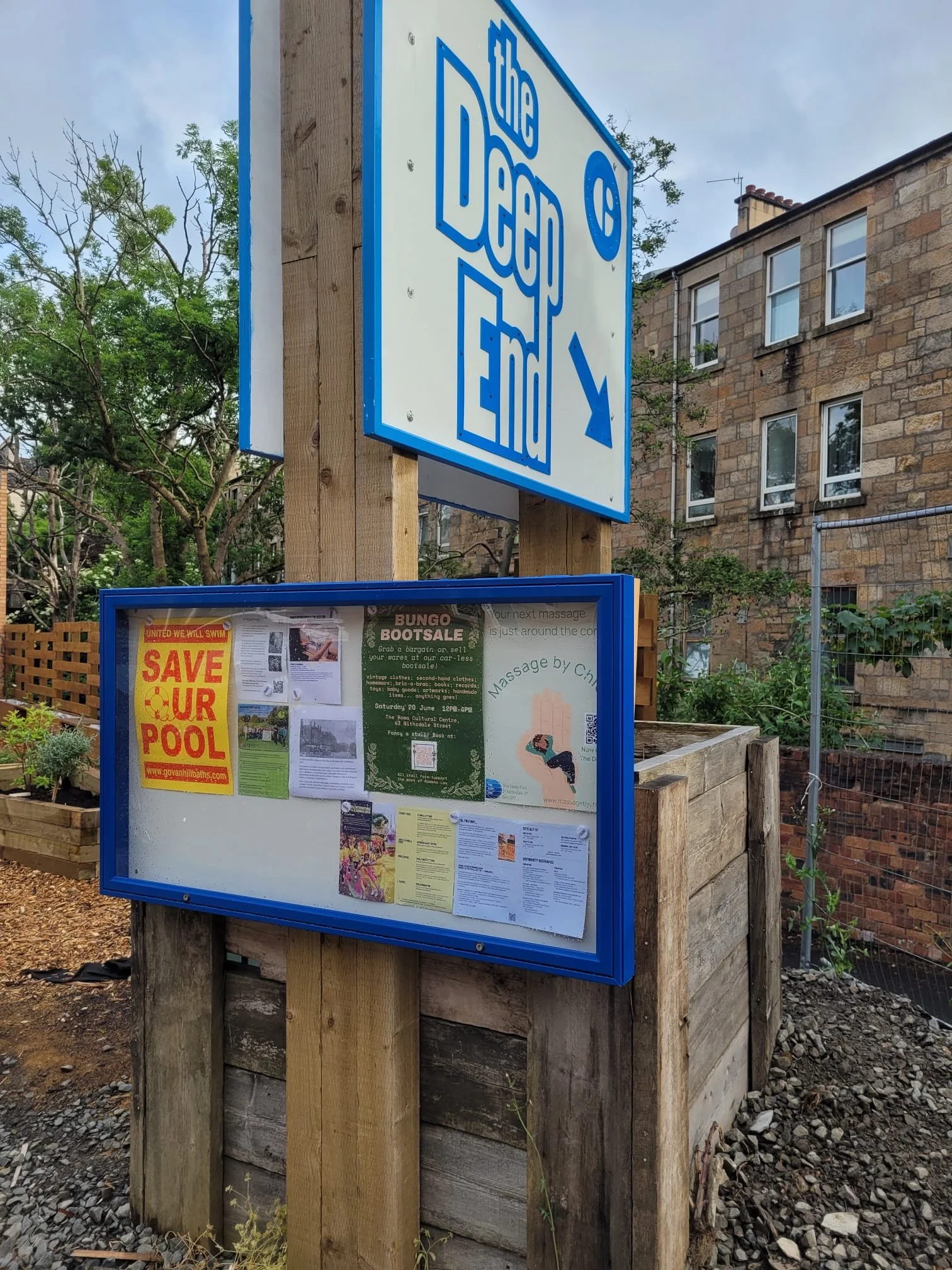 Public notice board with flyers and posters, located outdoors in front of a building with brick walls and windows, surrounded by trees and a wooden fence.