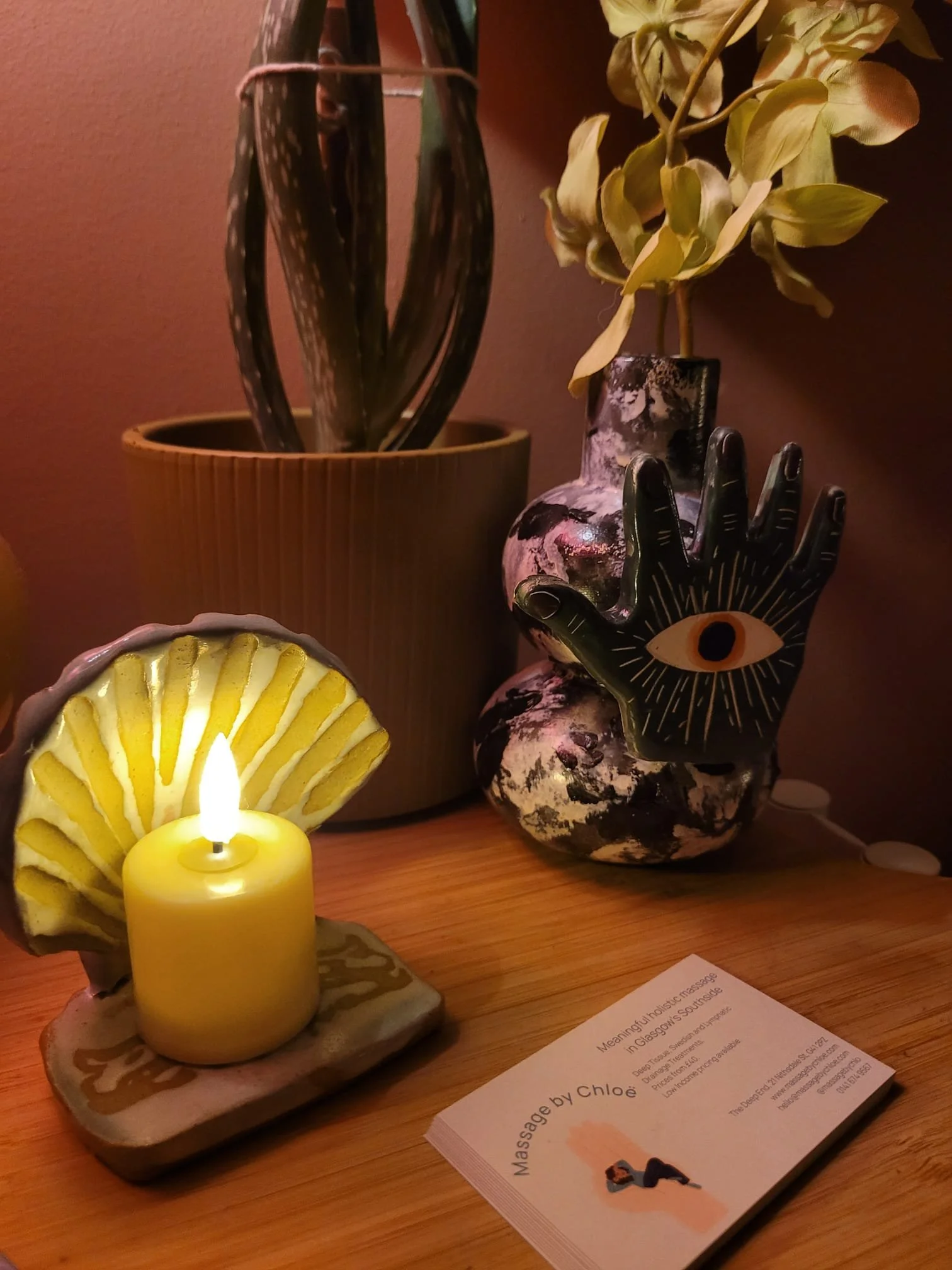 A lit yellow candle in front of a shell-shaped decorative dish, with dried flowers and a black and white vase with an eye design, all on a wooden surface.