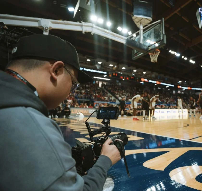 A person with a cap and glasses using a camera mounted on a stabilizer during a basketball game in an indoor arena filled with spectators.