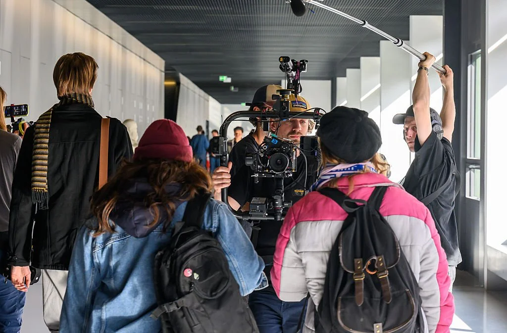 Film crew setting up a shot in a modern indoor location with large windows, including a camera operator, a person holding a boom microphone, and crew members with backpacks and jackets.