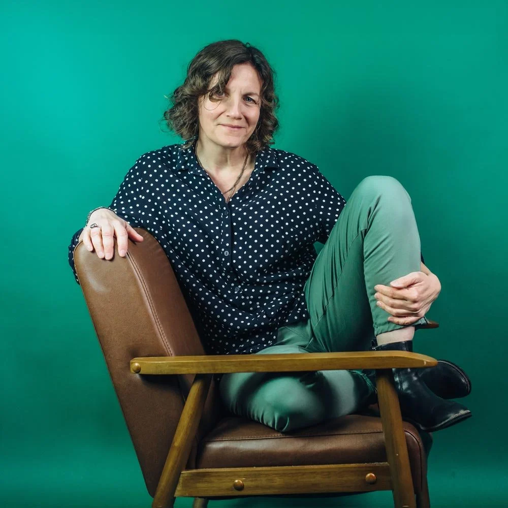 Nina Groop, Editor, Grant Writer, Fundraiser, Writer & Noticer. She has curly brown hair wearing a navy blue polka dot shirt, green pants, and black boots sitting relaxed in a brown leather and wooden chair against a green background.