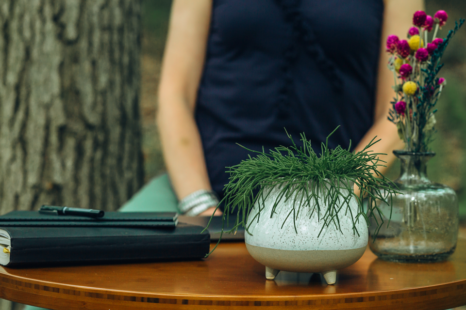 Close-up of a table with a potted plant, a notebook, and a glass jar with flowers, with a person standing behind it.