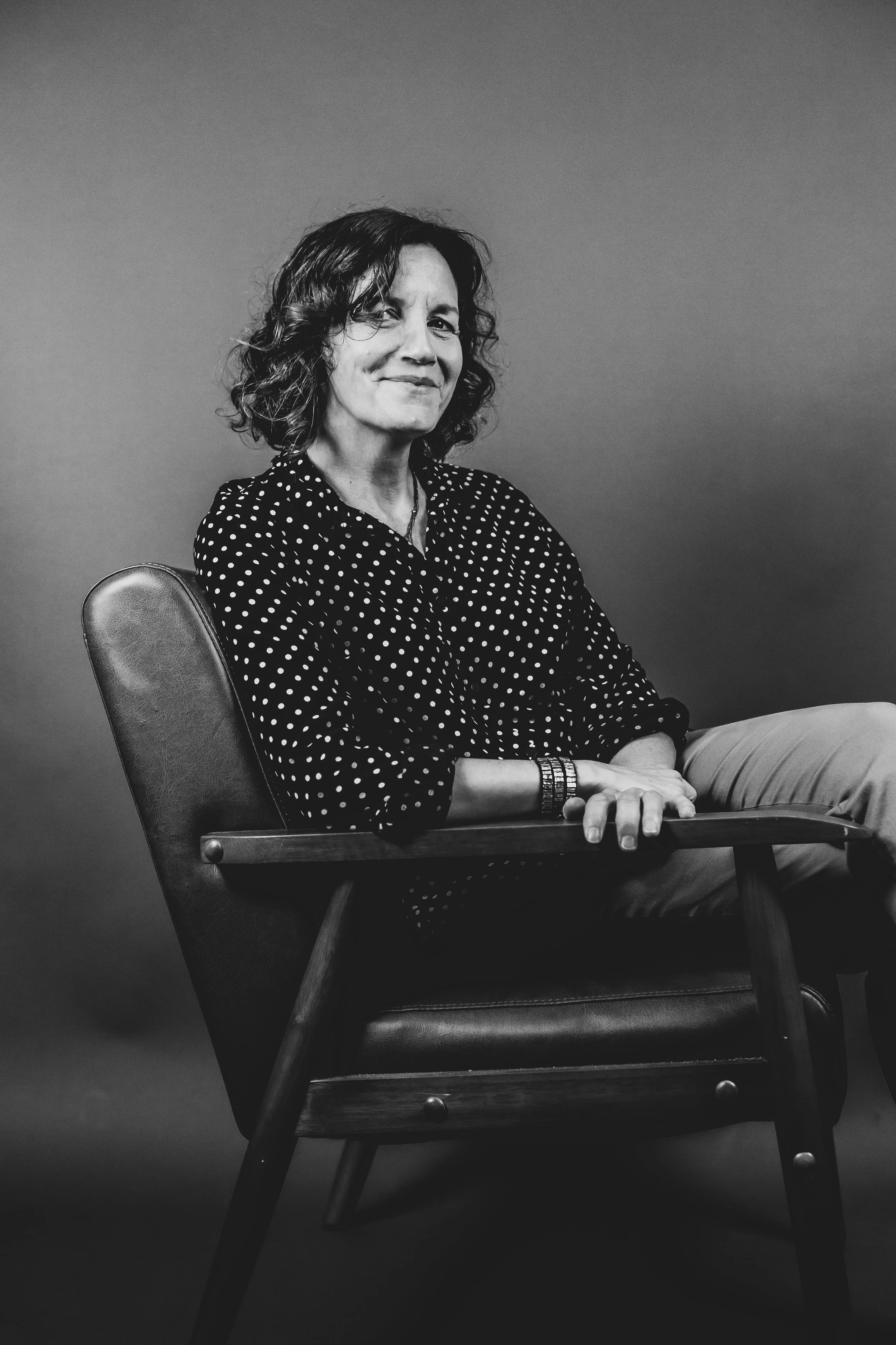 Nina Groop: Black and white photo of her sitting in a wooden chair, wearing a polka dot blouse, looking at the camera with a slight smile, against a plain background.