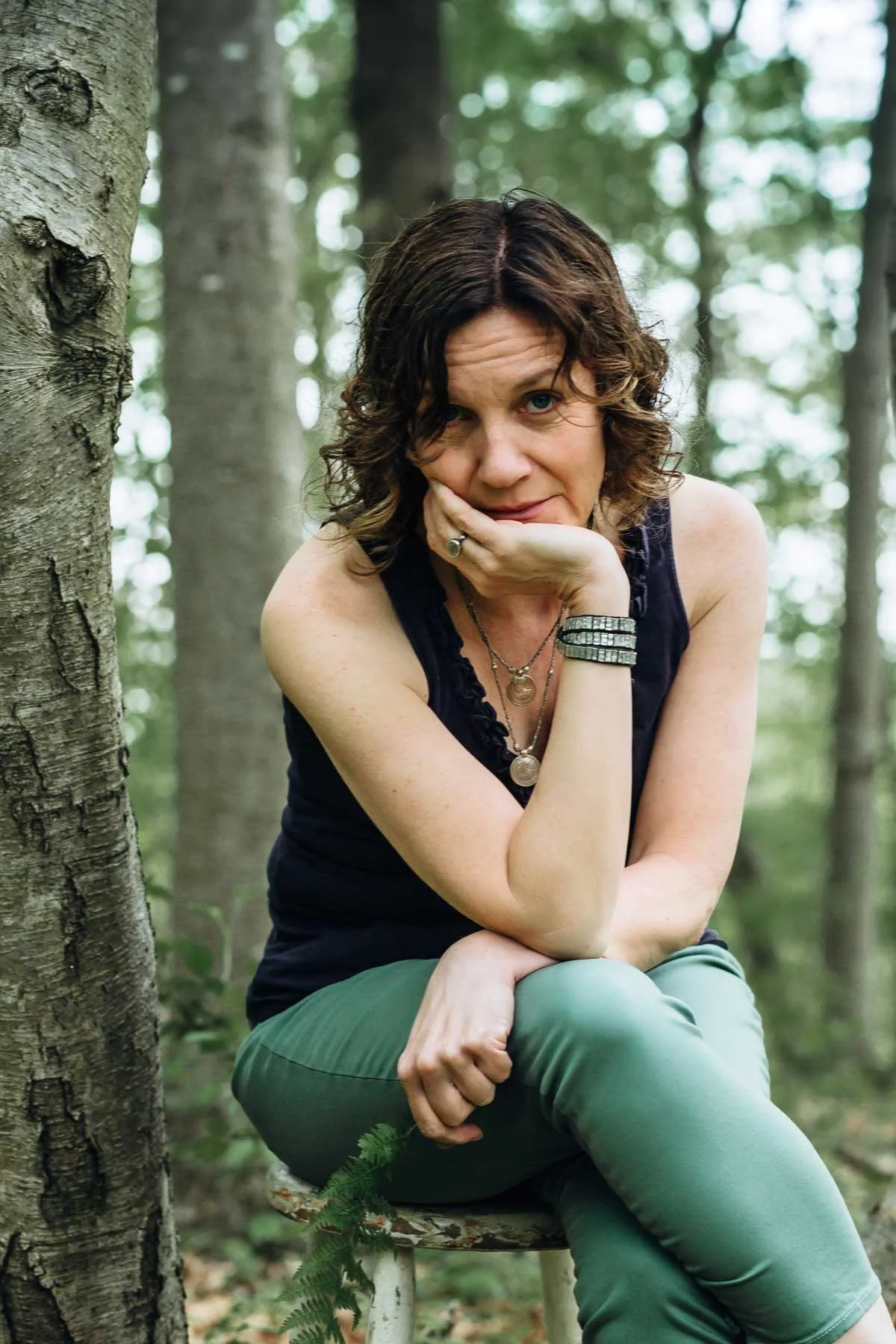 Nina Groop, writer, noticer: a woman with curly brown hair and blue eyes sitting on a tree stump in a forest, resting her chin on her hand and looking at the camera.