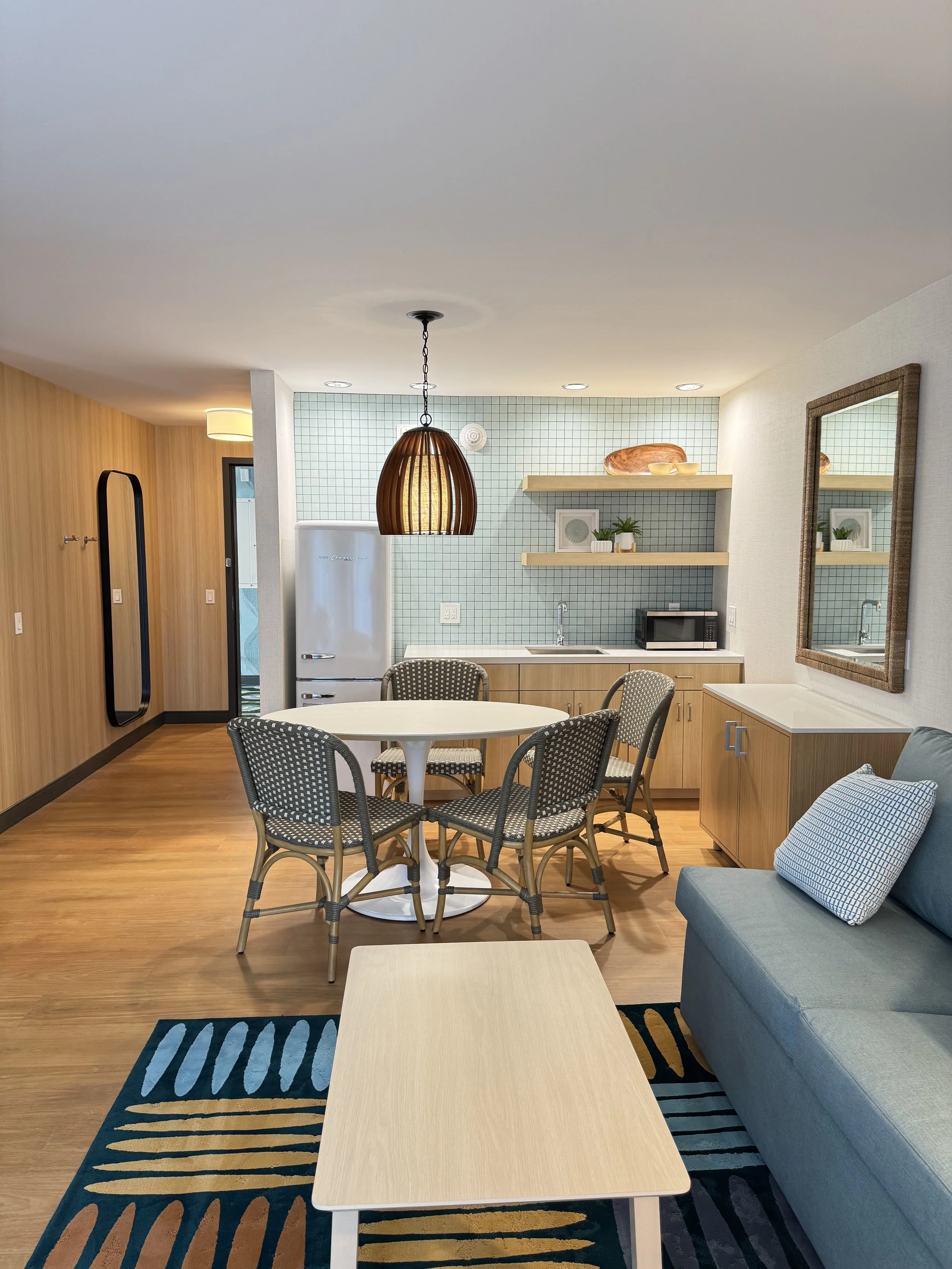 Living room with wooden floor, blue and beige rug, blue sofa with striped pillow, round white table with four wicker chairs, a black-framed mirror, light wood cabinets, a small kitchenette with a white refrigerator, a microwave, two floating shelves with decorative items, and a hanging wooden pendant light.