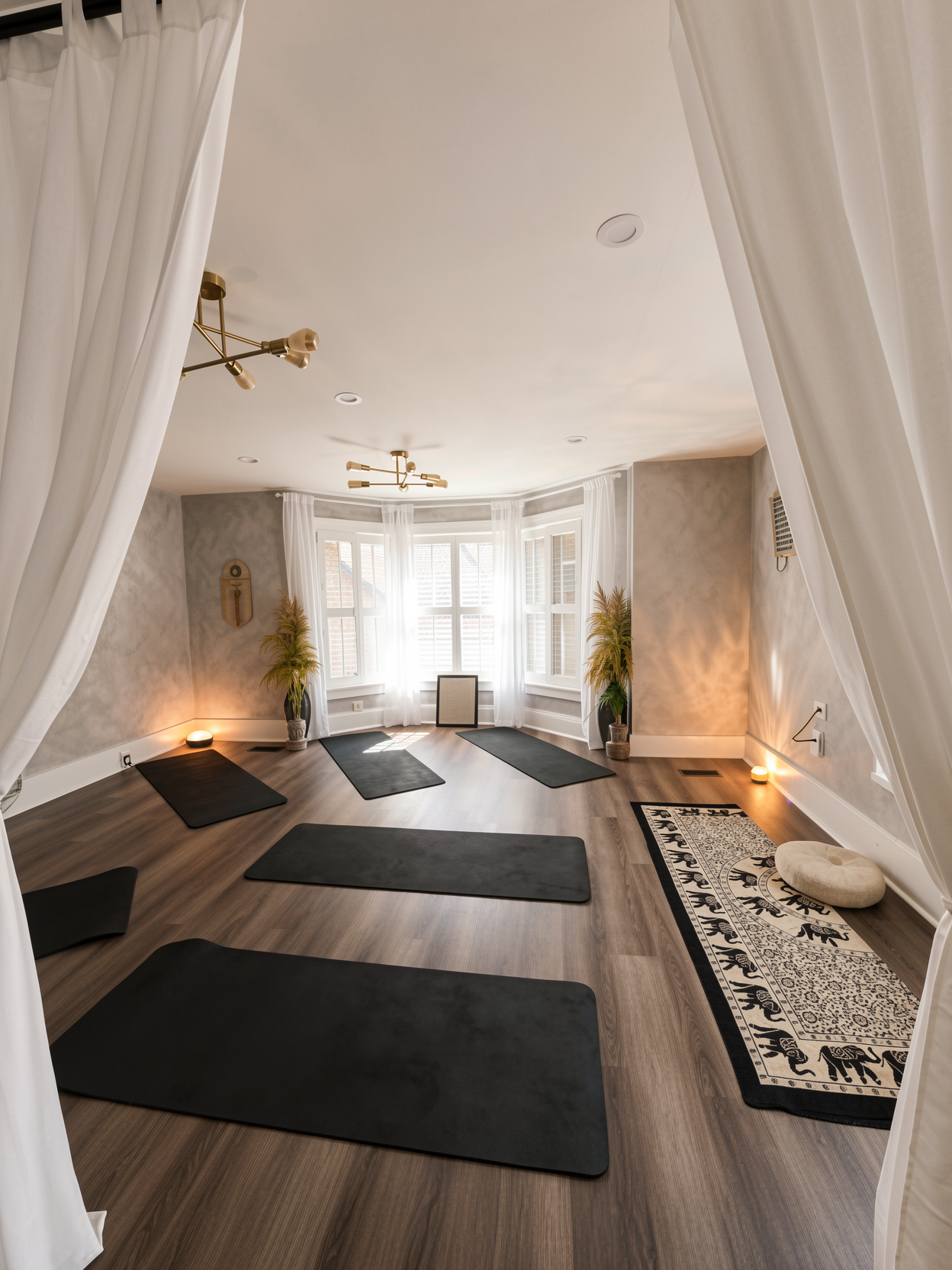A cozy yoga or meditation room with five black yoga mats on a wood floor, white curtains, plants, soft lighting, and a patterned rug with a round cushion.