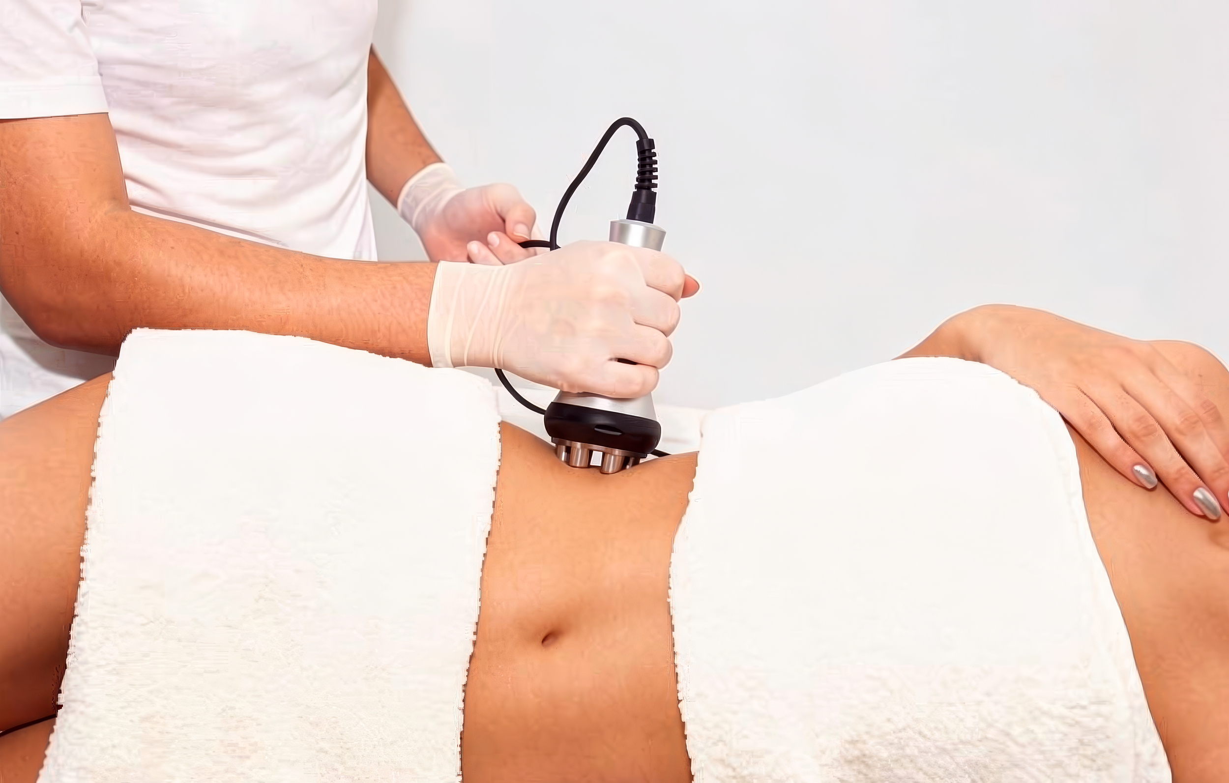 Medical professional performing a procedure on a patient using a handheld device with a cord, patient lying on an examination table with their arm extended.