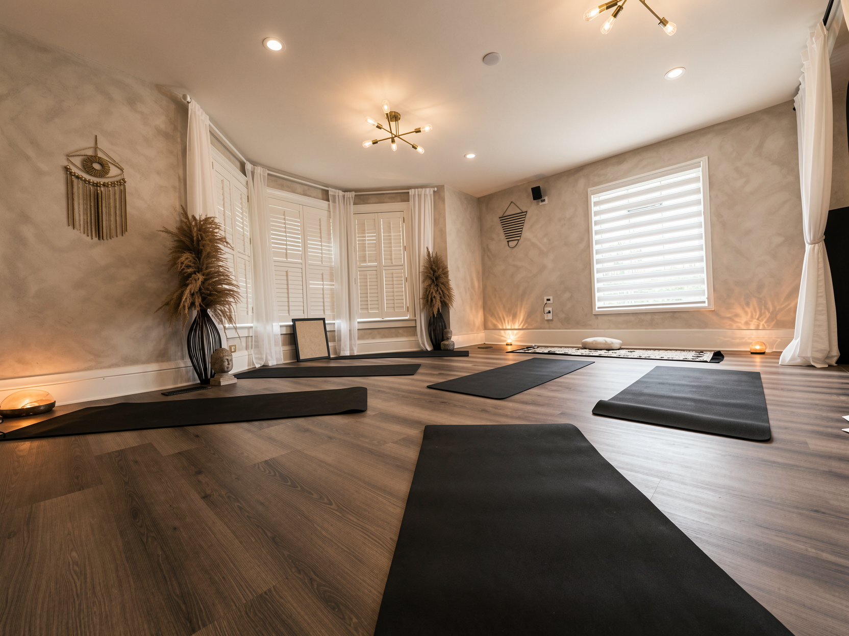 A peaceful yoga studio with black mats on wooden floors, white curtains, dried plants, decorative wall art, and warm lighting.