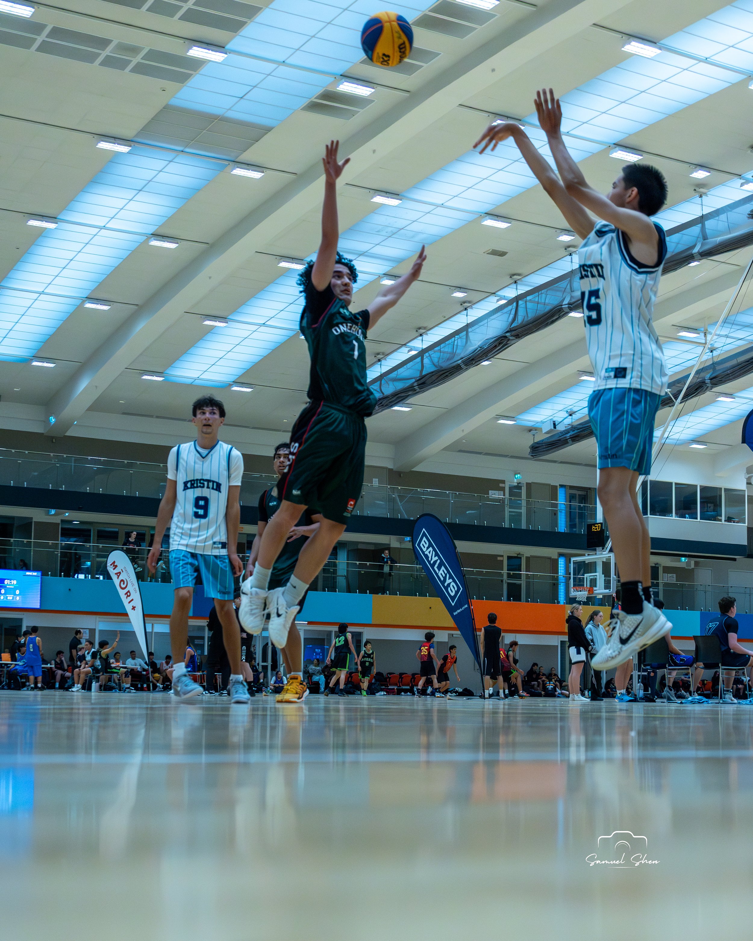 Young athletes playing basketball in an indoor gymnasium with spectators and banners around.