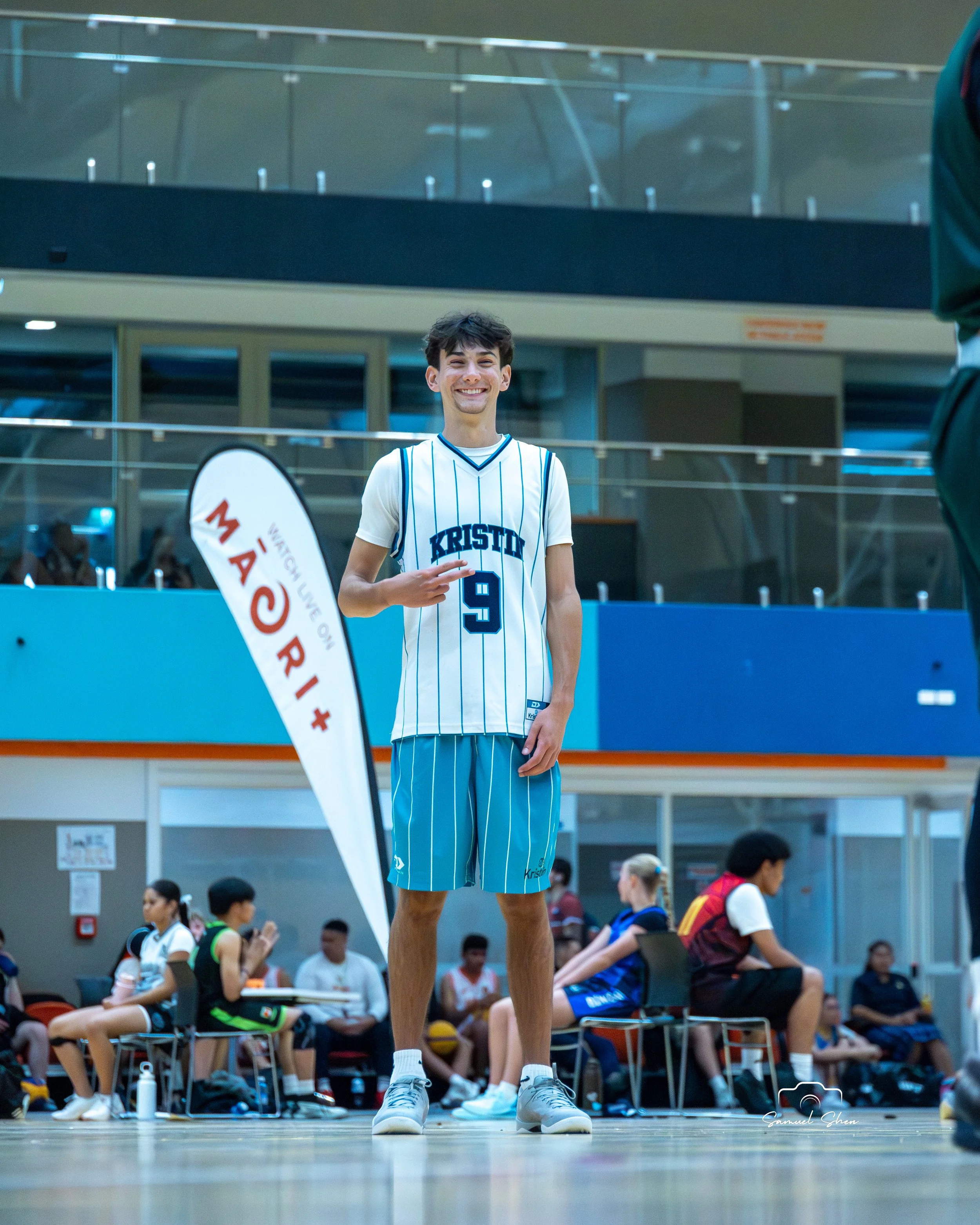 A young male basketball player wearing a white and blue striped basketball uniform with the number 9 and the word 'KRISTO' on the front, standing on a basketball court with a smile, in an indoor sports arena filled with people sitting on benches and 