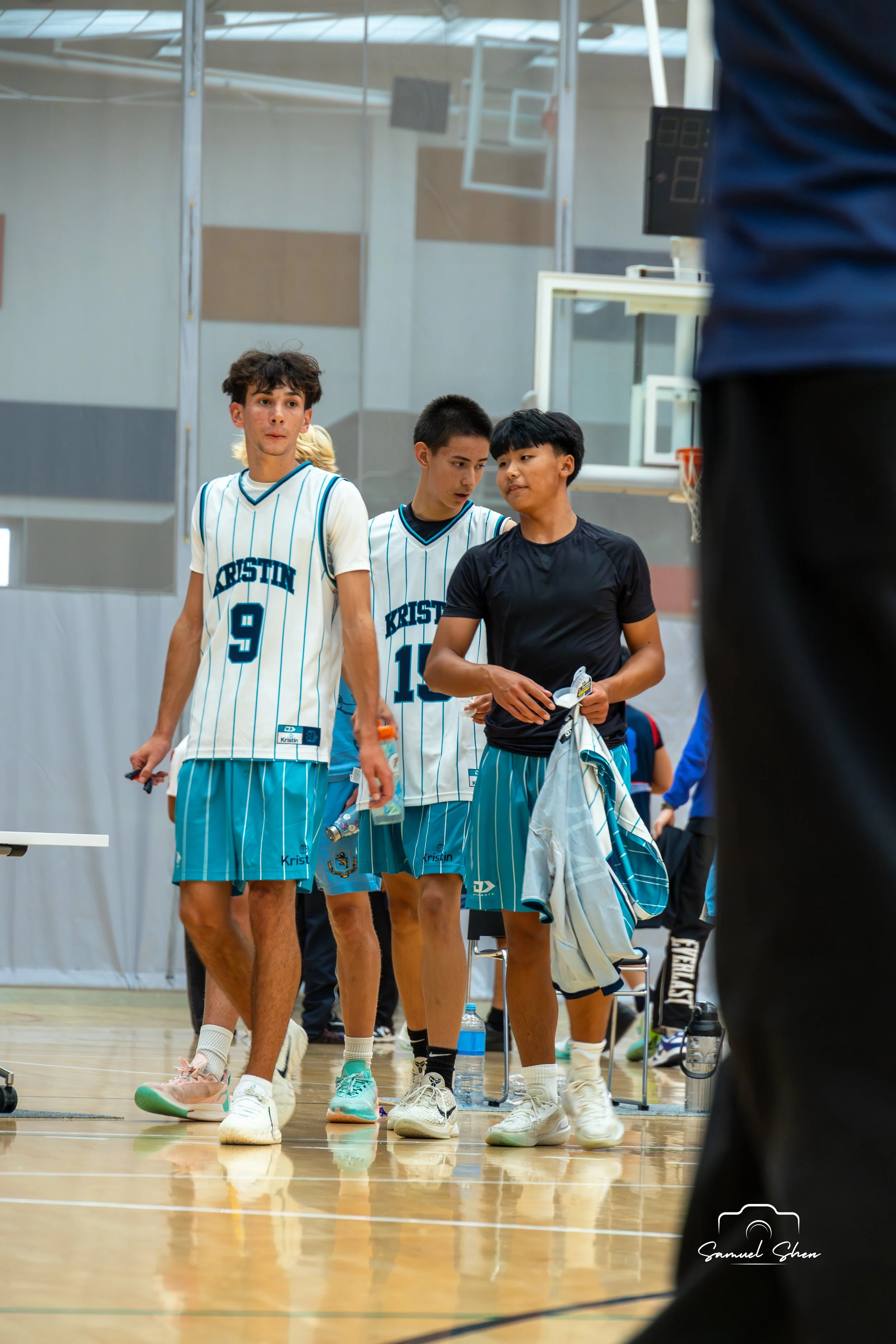 Young male basketball players wearing striped jerseys walk in a gymnasium, carrying sports equipment and water bottles, with a basketball hoop and digital scoreboard in the background.