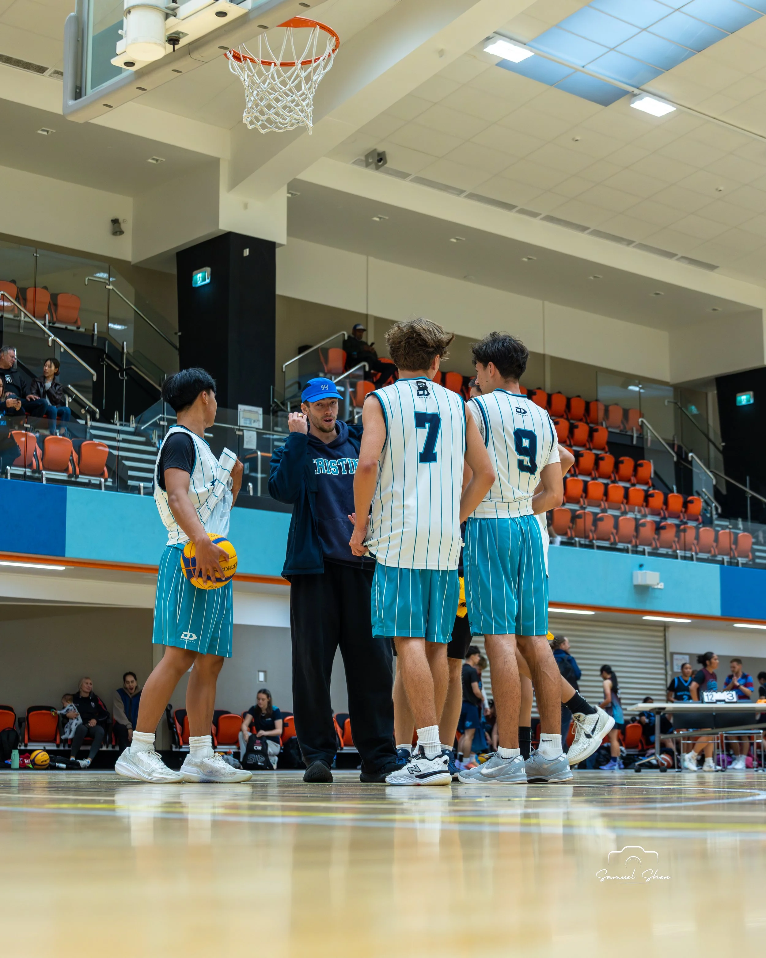 A group of basketball players in teal and white uniforms standing and talking on the court, with some people sitting on orange seats in the background of an indoor gym.