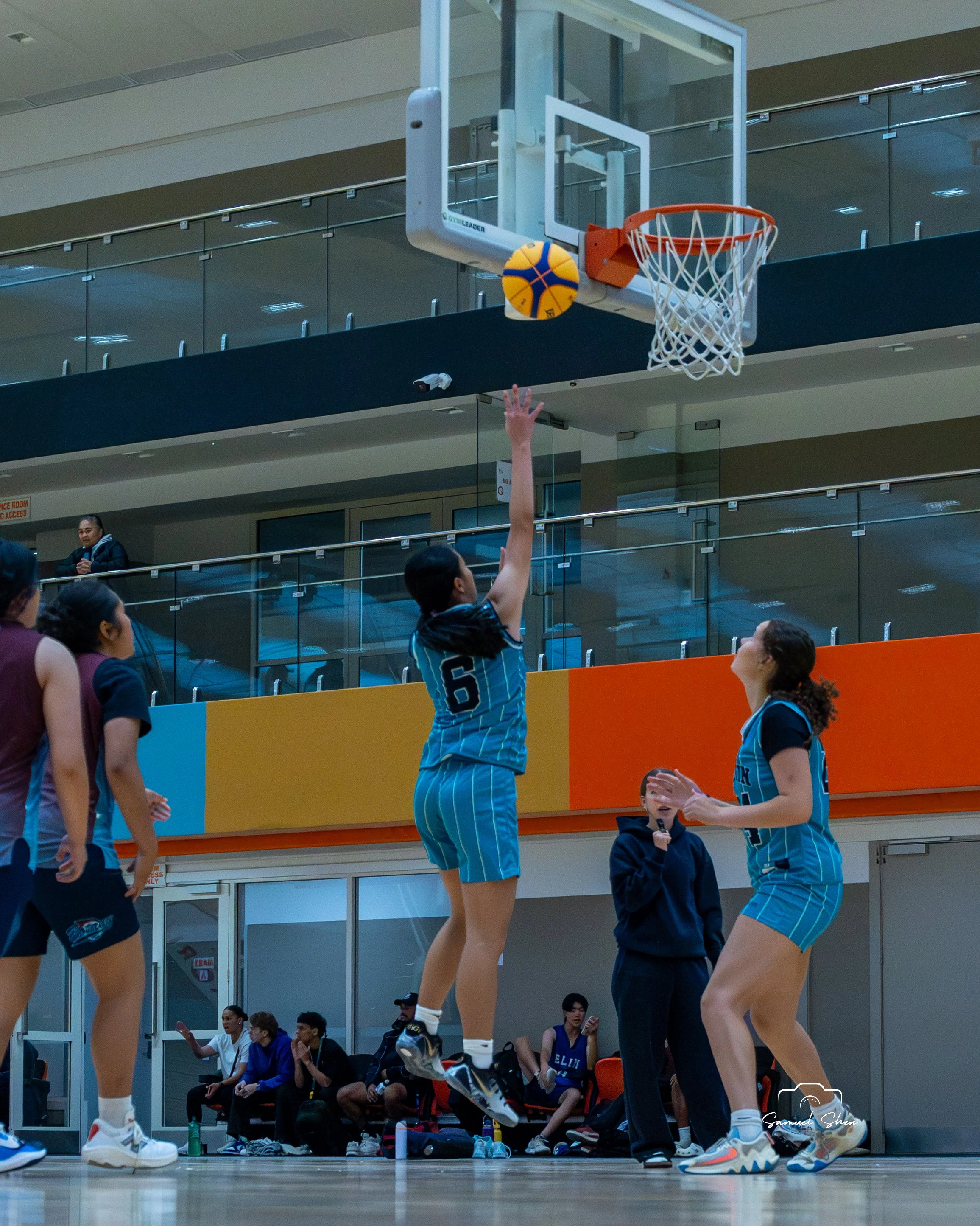 Young female volleyball players in teal uniforms practicing warm-up, with one jumping to hit a volleyball near the net. Other players and coaches observe while seated on benches inside a sports facility.