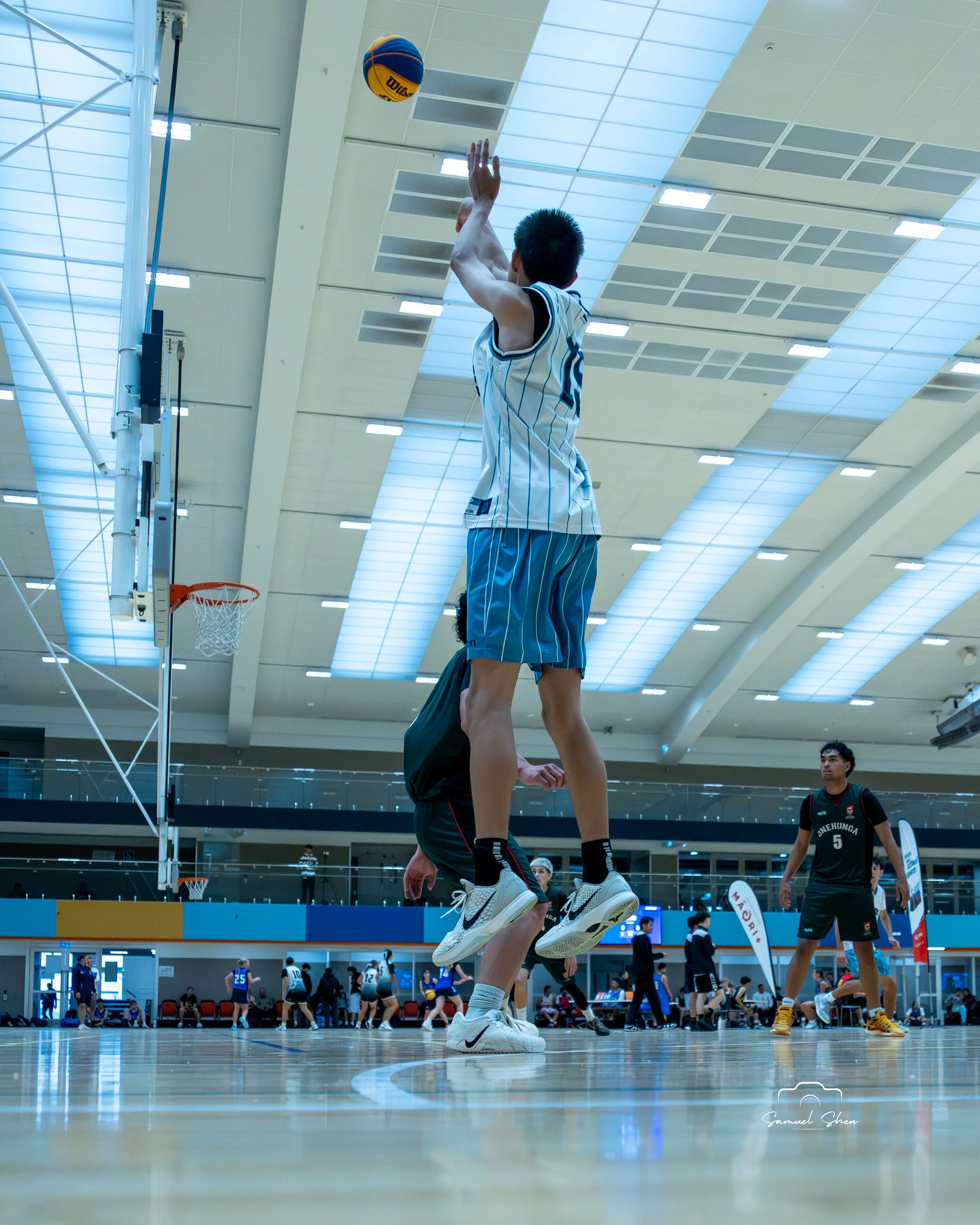 A volleyball player in a white and blue uniform is jumping to block a volleyball during a game inside a sports hall. Other players and spectators are visible in the background.