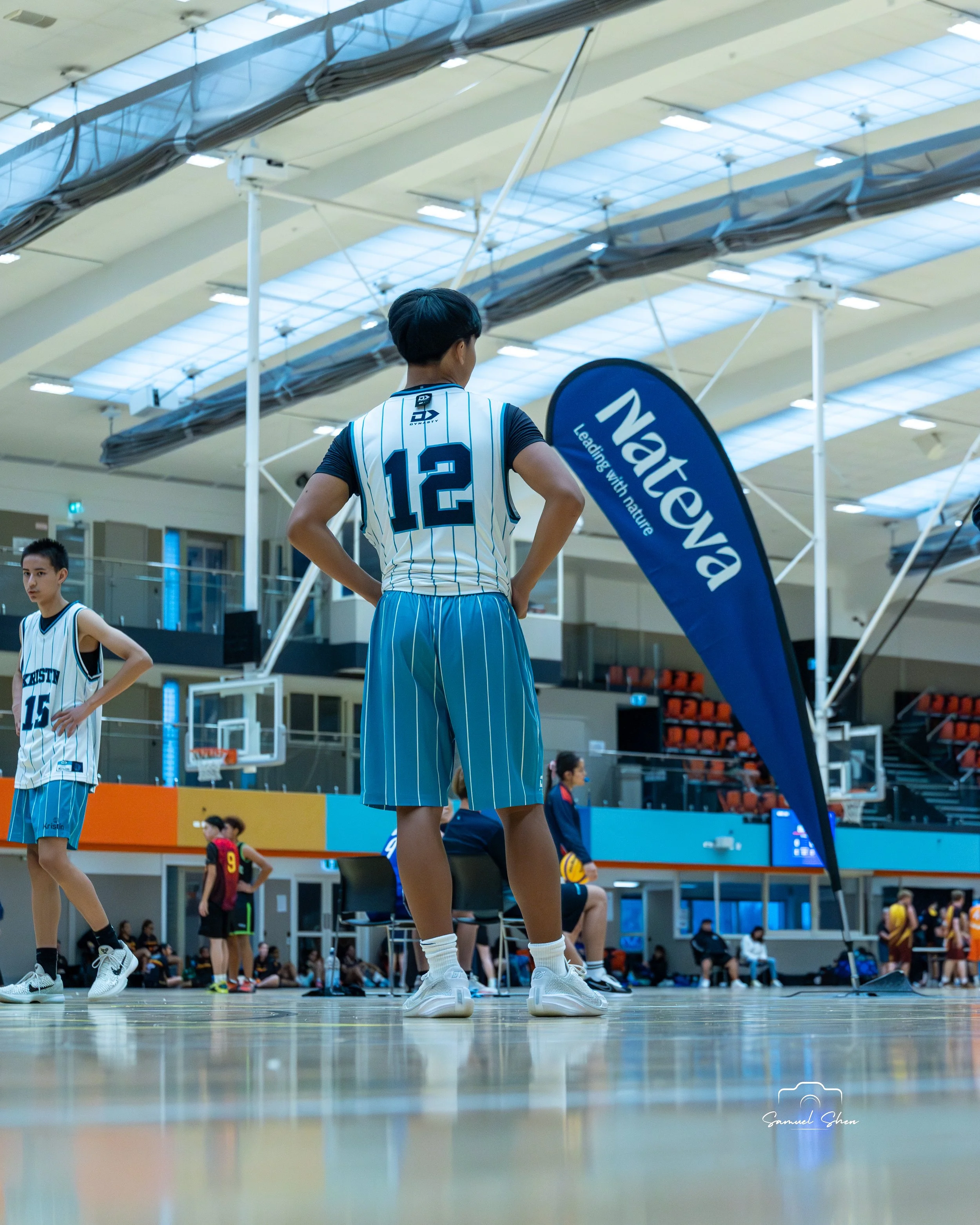 A female basketball player wearing a white and blue striped uniform with the number 12 on her back stands on an indoor court with others around, some seated and some playing, and large banners next to basketball hoops in the background.