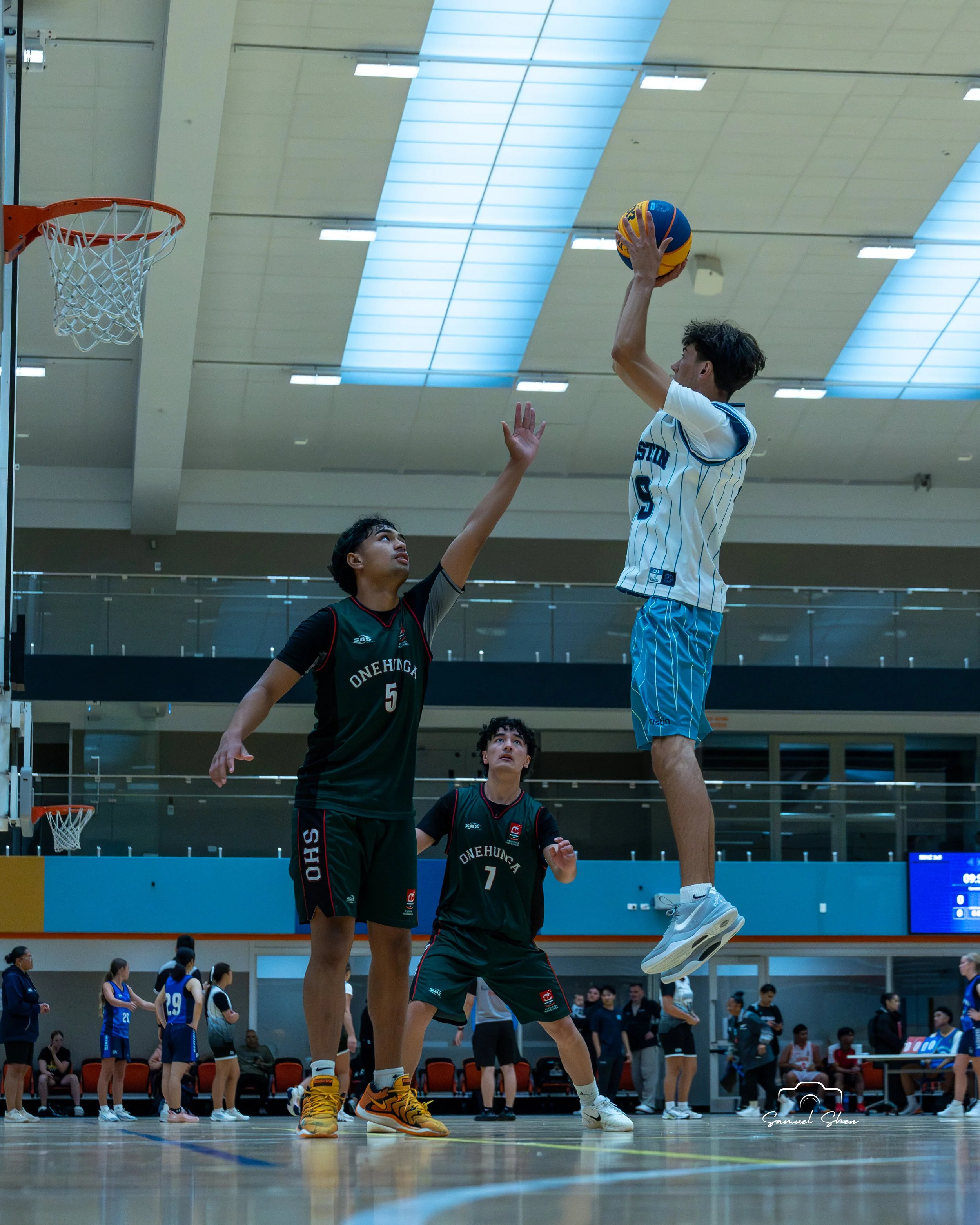 A young male basketball player wearing a white and blue jersey jumps to shoot with a colorful basketball, while another player in a black and green jersey defends, on an indoor court with spectators and other players in the background.