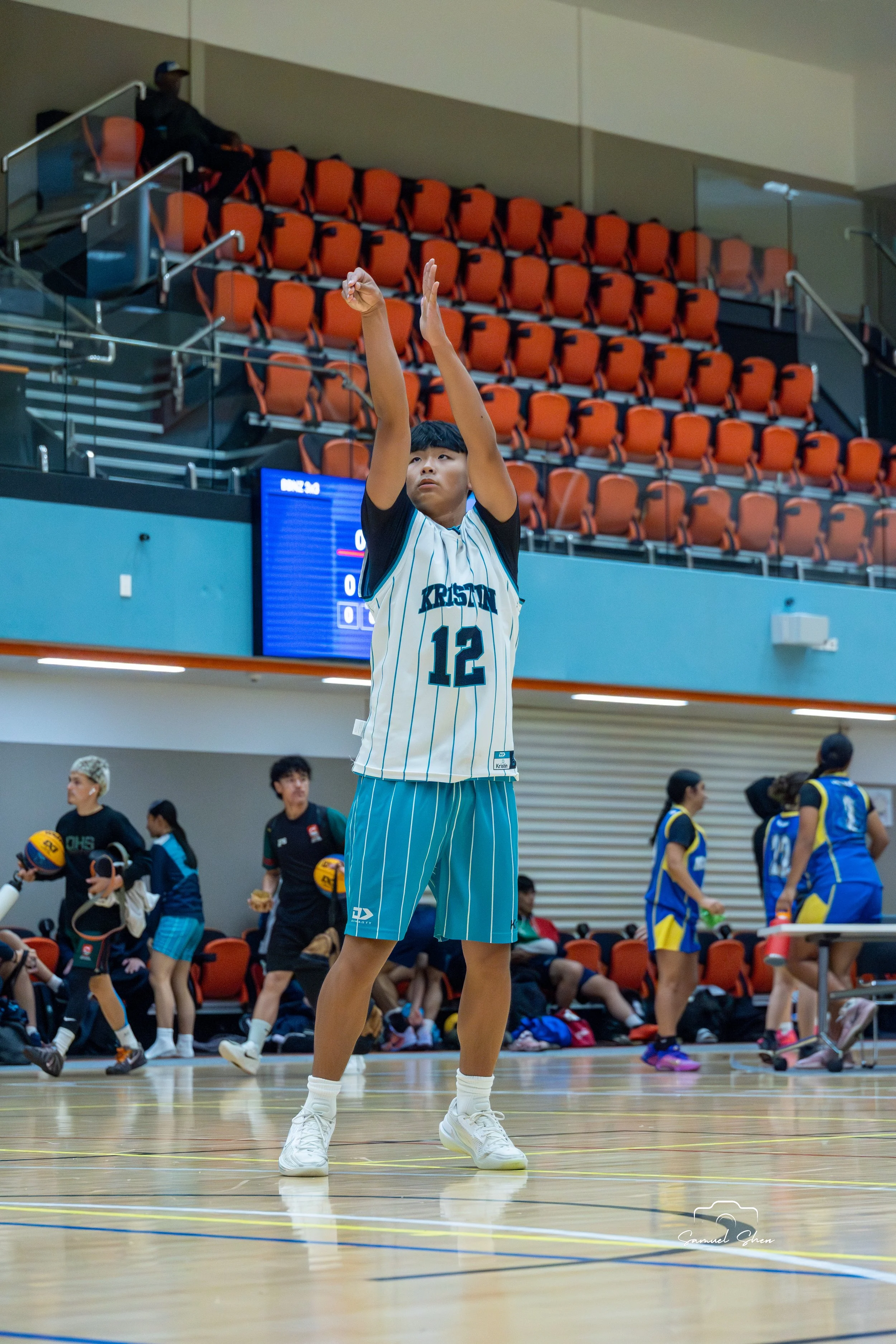 A young boy on an indoor basketball court, wearing a sports jersey with the number 12, white sneakers, and lying on his back with arms raised.