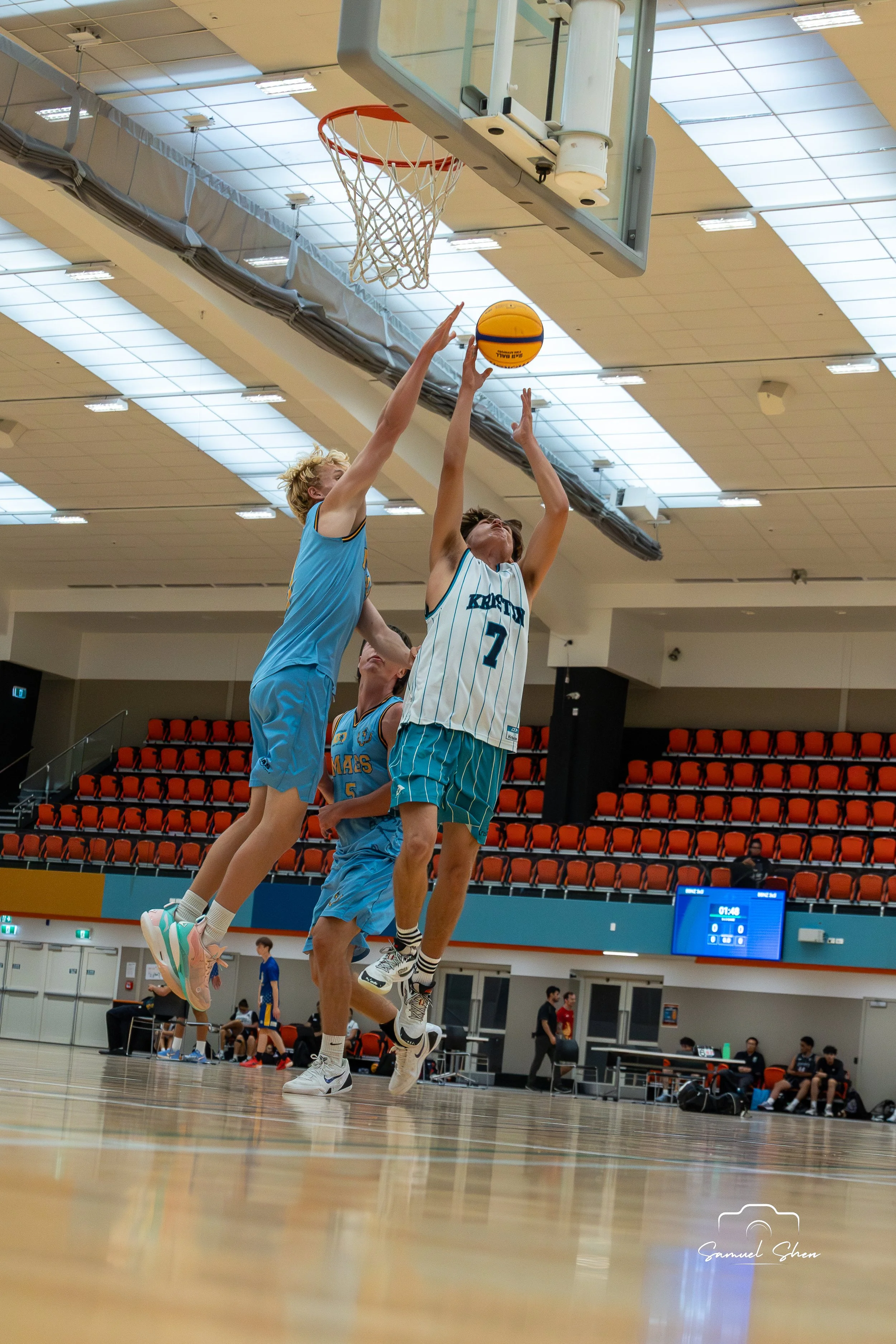 Two basketball players in mid-air jump for a basketball near the hoop during a game in a gymnasium, with other players and officials visible in the background.
