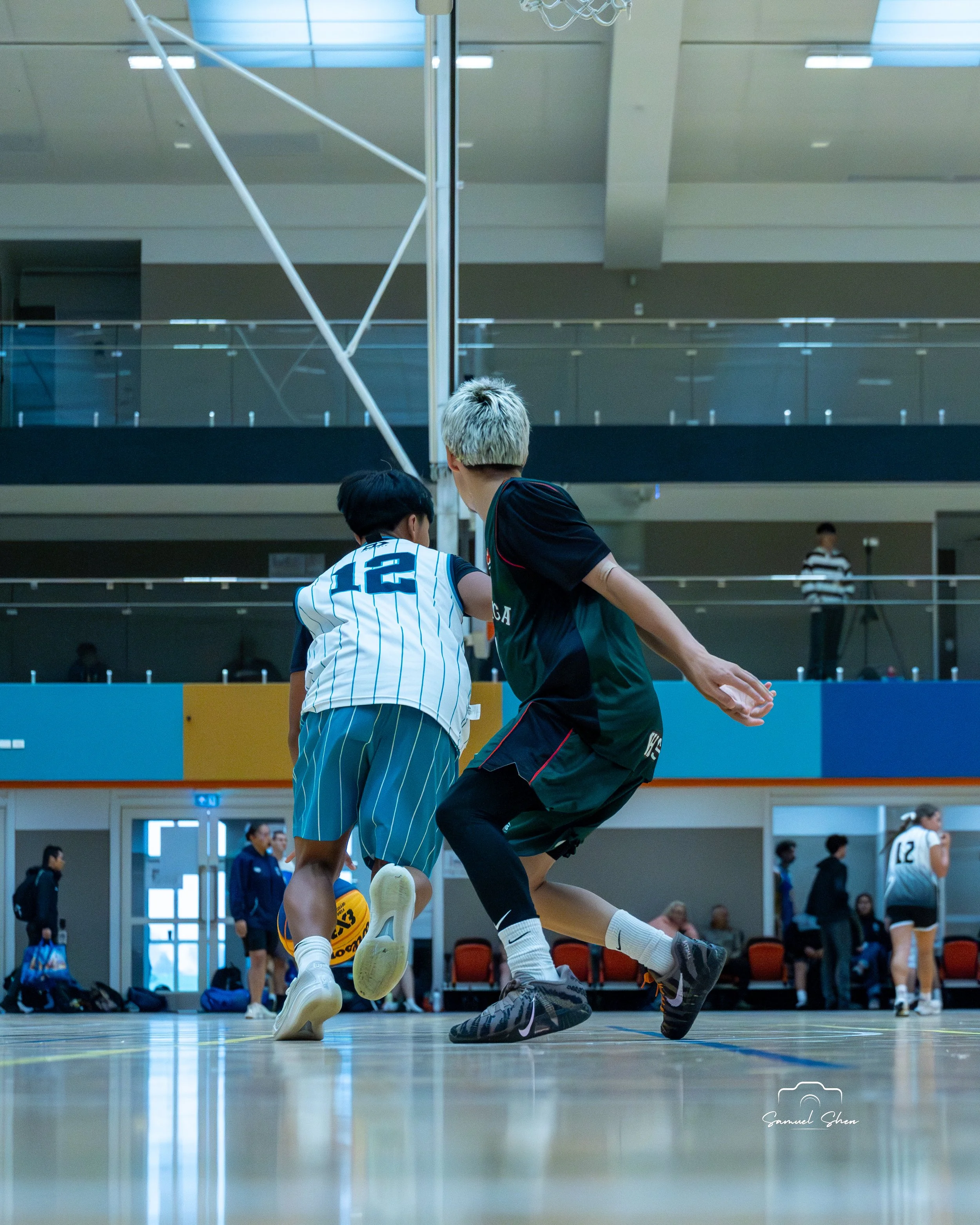 Two young men playing basketball inside an indoor court. One is wearing a white and blue striped jersey with the number 12, and the other is in black athletic clothing. They are jumping and competing for the ball near the hoop, with spectators and st