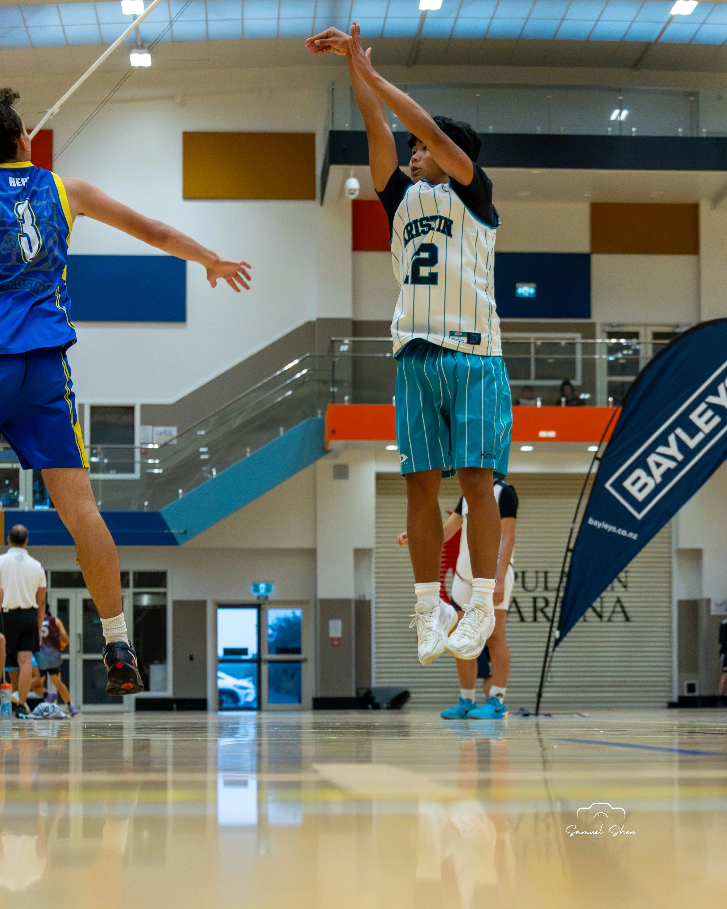 Two basketball players are jumping to reach the ball during a game in an indoor stadium; one player is wearing a white jersey with blue pinstripes and the number 2, while the other is wearing a blue jersey with yellow accents.