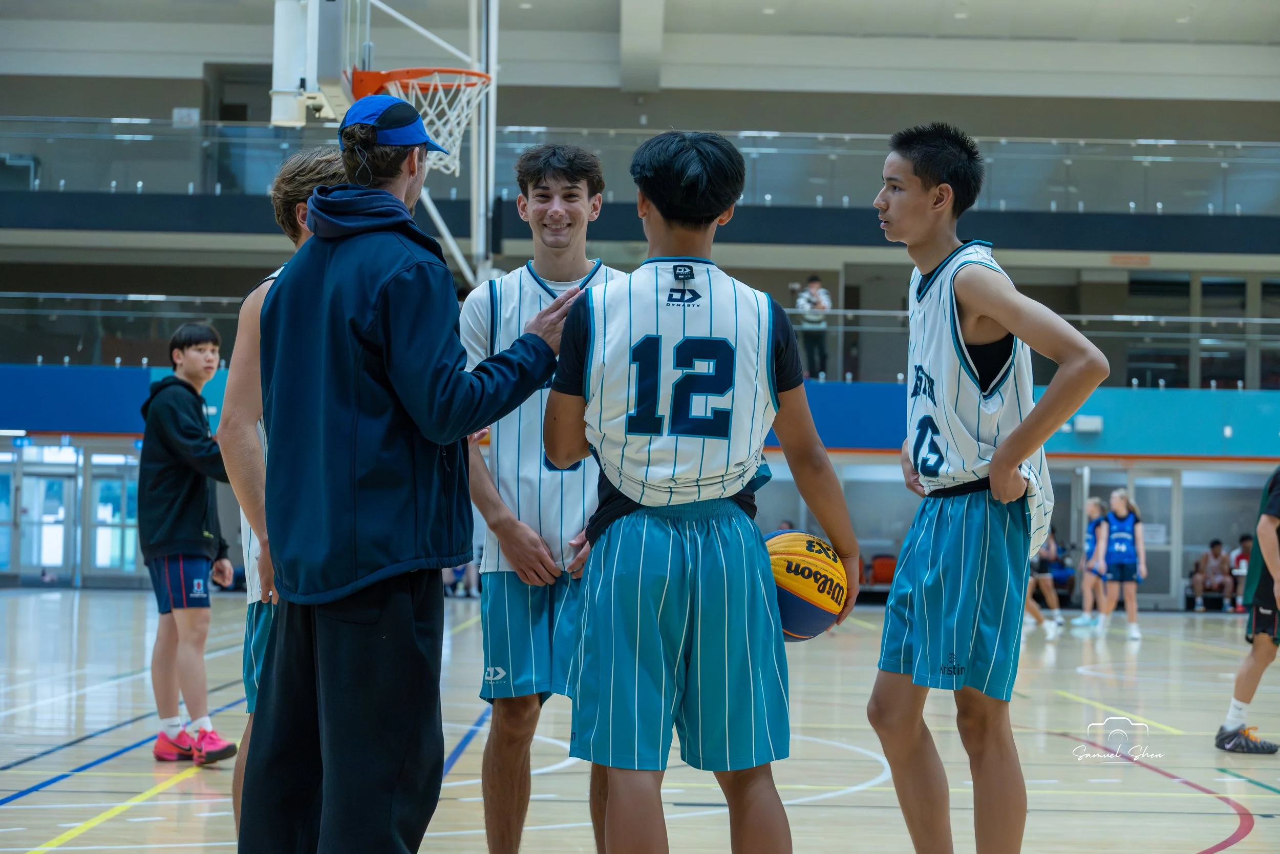 A group of young basketball players and their coach having a discussion on the court in an indoor gymnasium. The coach is talking to the players, who are in basketball uniforms, with one holding a basketball.