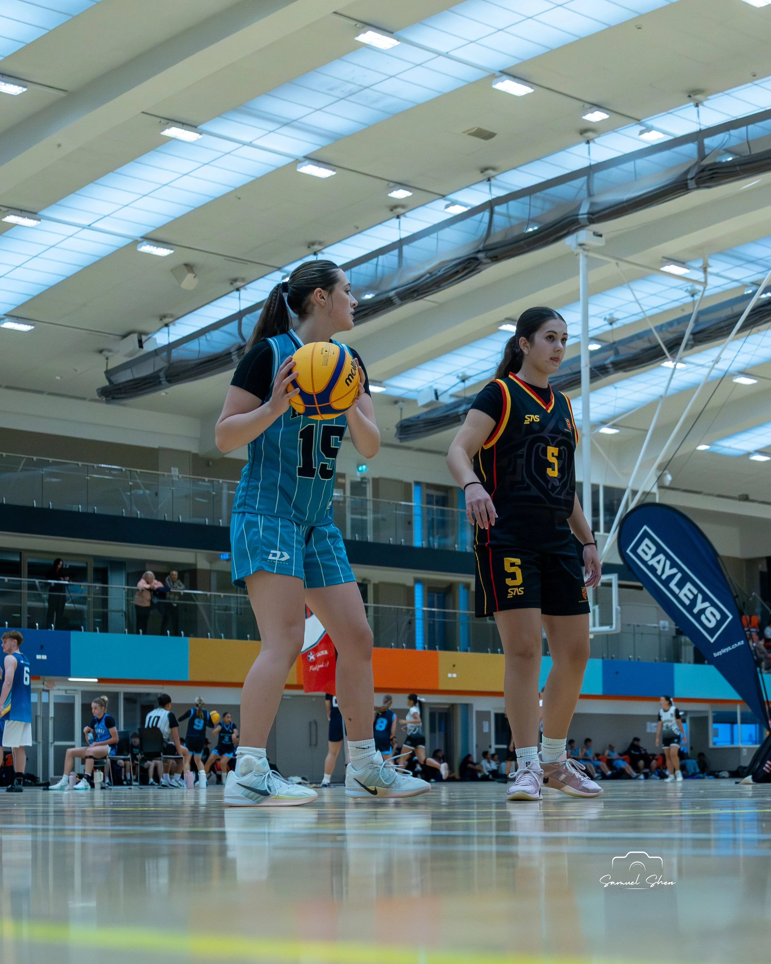 Two female basketball players on the court, one holding a basketball and wearing a blue uniform with the number 15, and the other wearing a black uniform with red and yellow accents and the number 5. They are in an indoor sports arena with other play