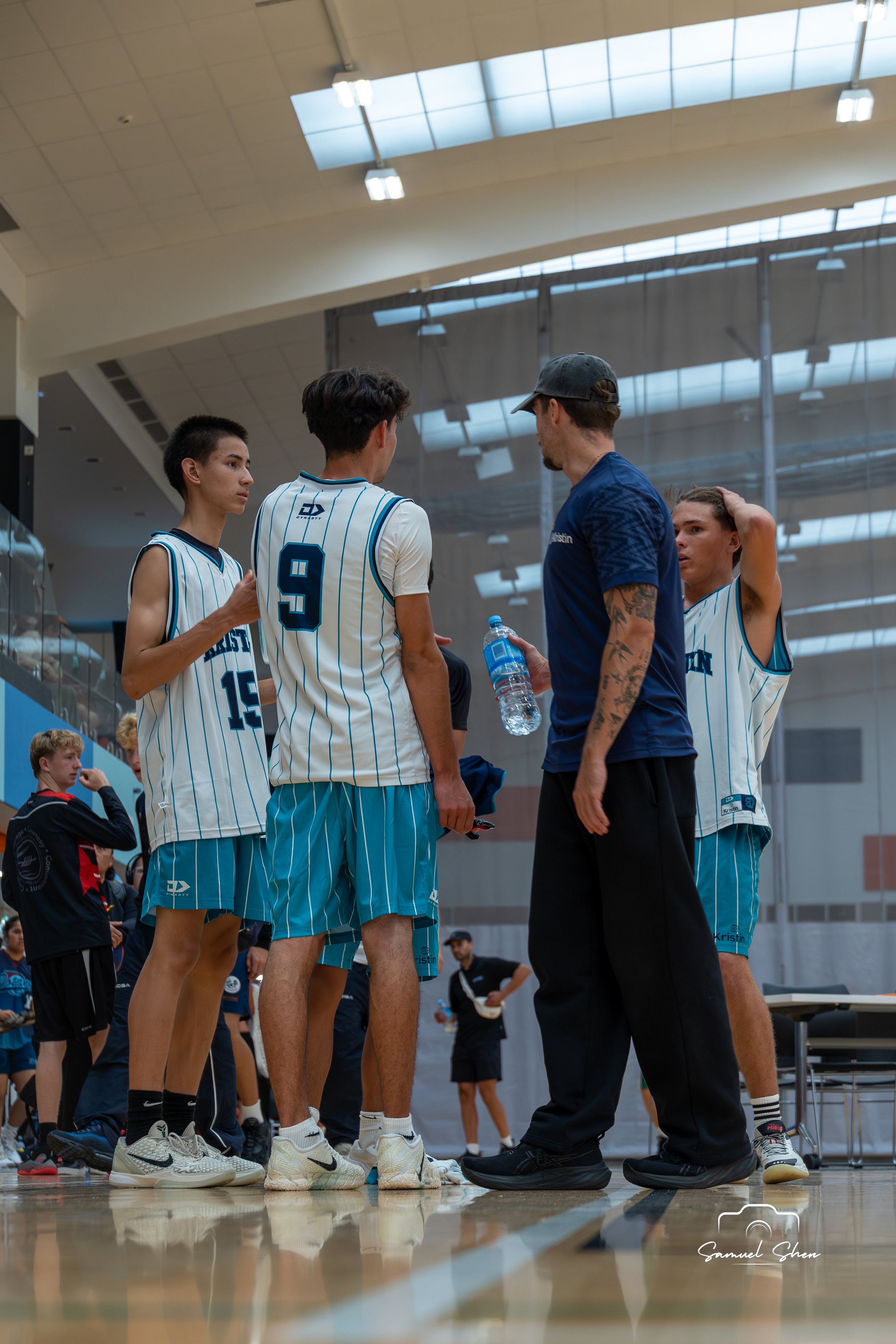 A basketball coach talking to four players during a timeout in an indoor basketball court. The players are wearing white and blue striped uniforms, and the coach is dressed in black with a cap. The players are listening attentively.