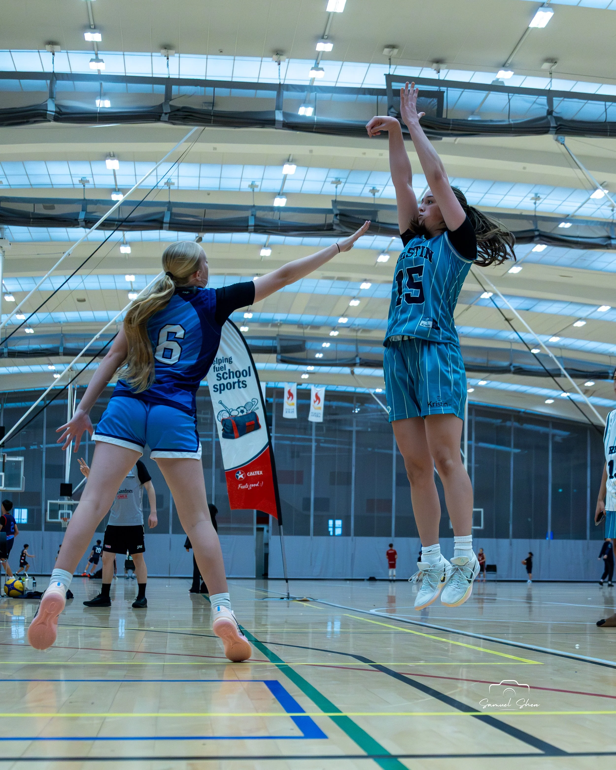 Two female basketball players jumping to block the ball during a volleyball game in an indoor gymnasium. The gym has a high ceiling with lights and a basketball hoop visible in the background.