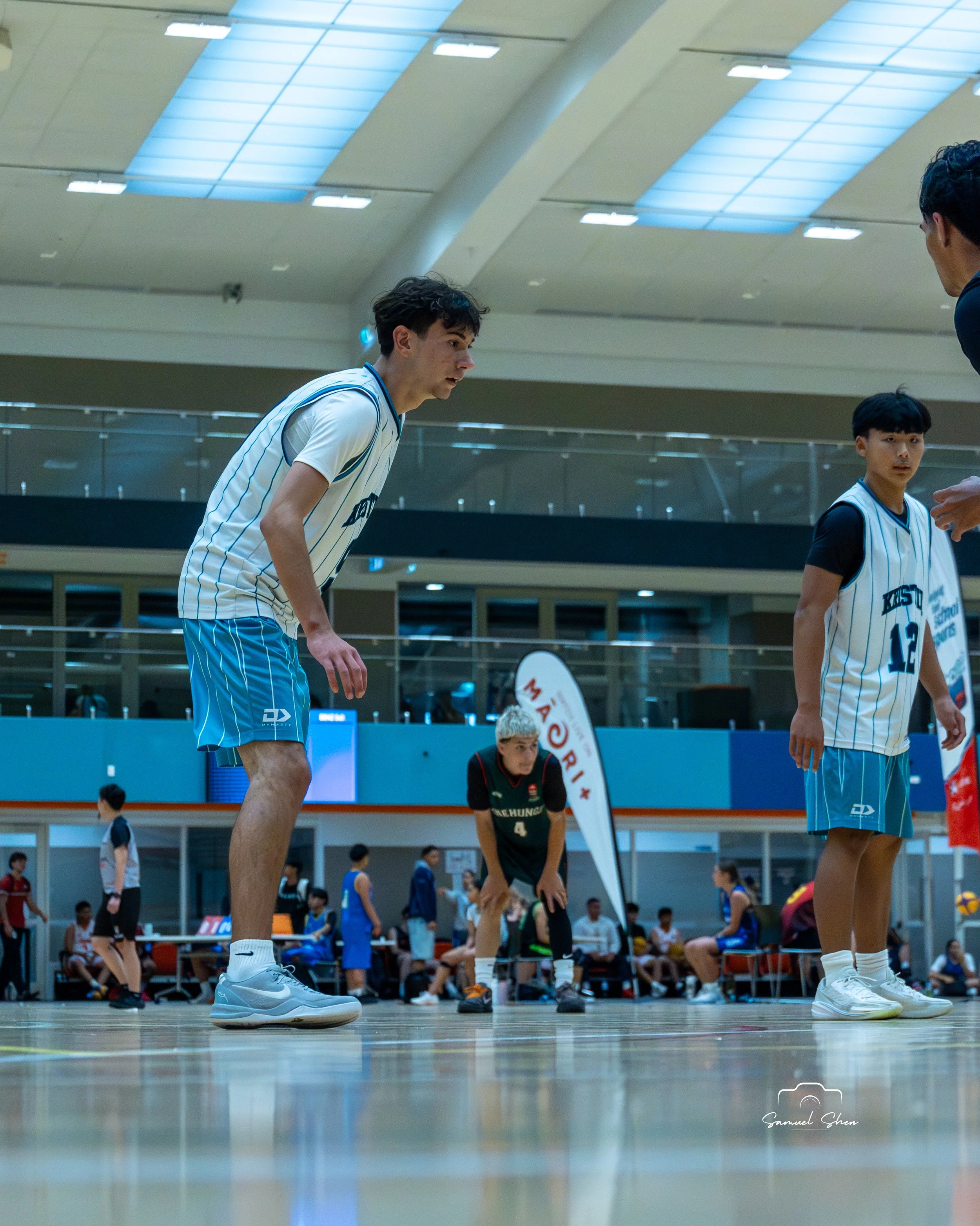 Interior view of a basketball court with players in uniform preparing for a game or practice, focusing on their shoes and lower bodies, under bright ceiling lights.
