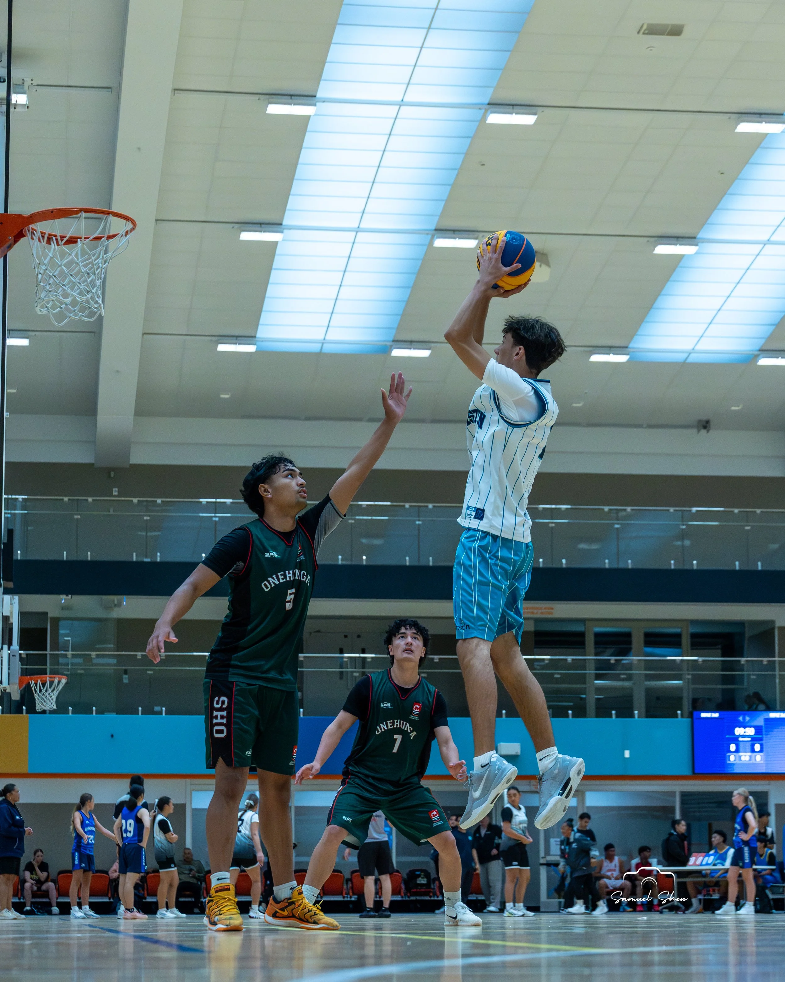 An indoor basketball game with players in action. One player in a white and blue uniform jumps to shoot the ball while another player in a black and red uniform tries to block the shot. Other players and spectators are visible in the background.