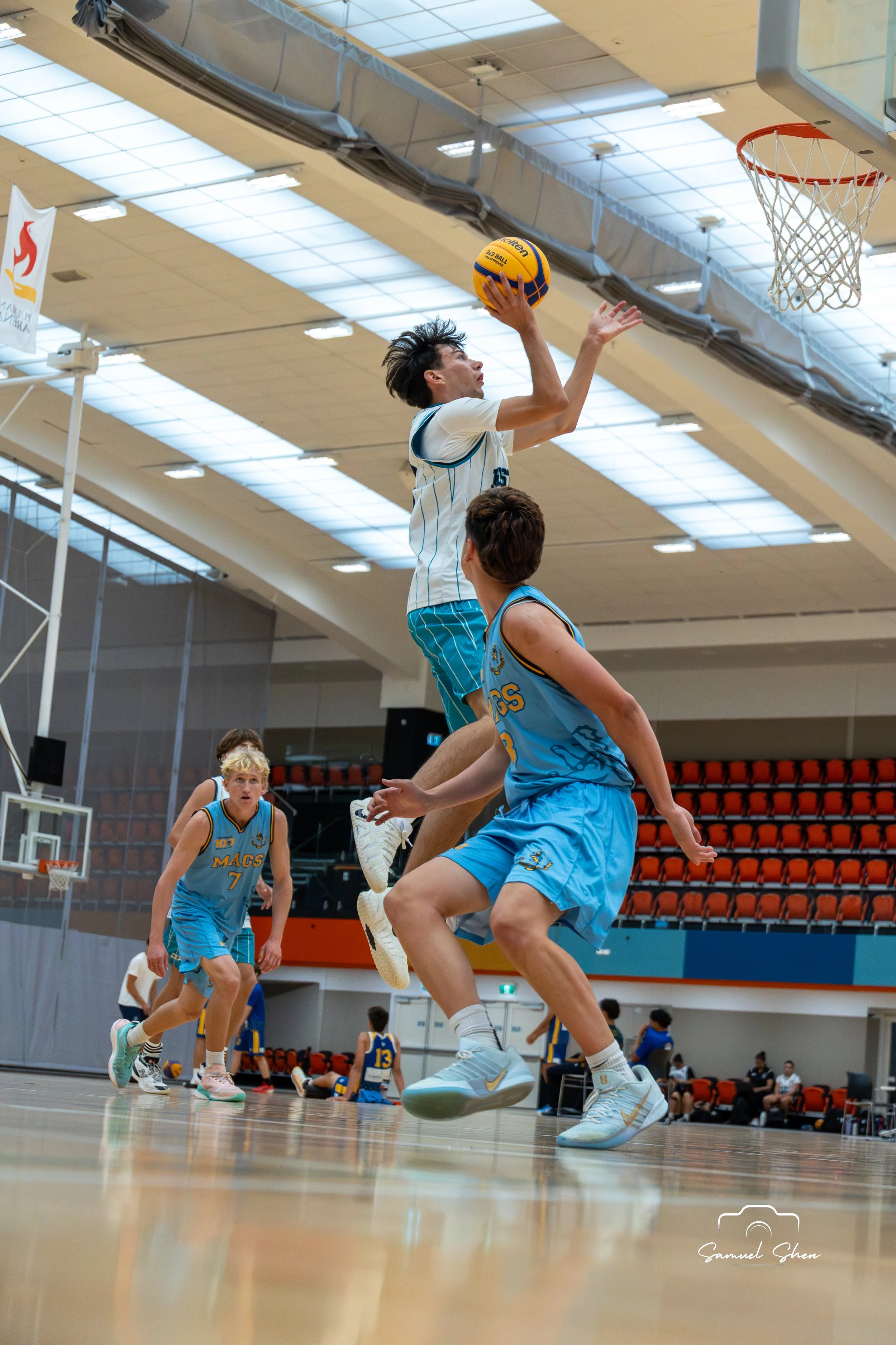 A basketball player jumping to make a shot in an indoor court. Other players and spectators watching in the background.