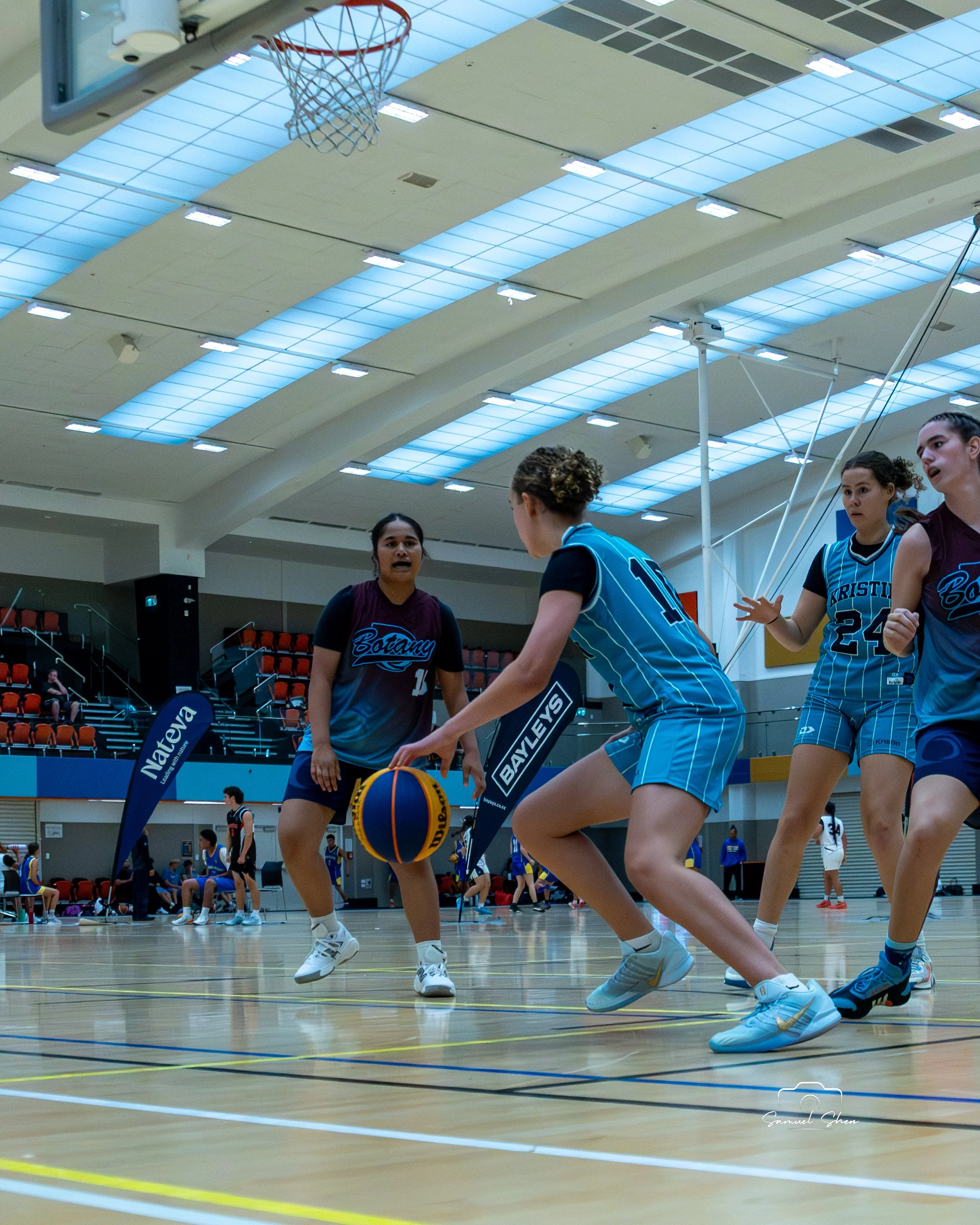 Women playing basketball in an indoor court, some players are wearing blue uniforms and others are in dark uniforms, with spectators sitting in the background.