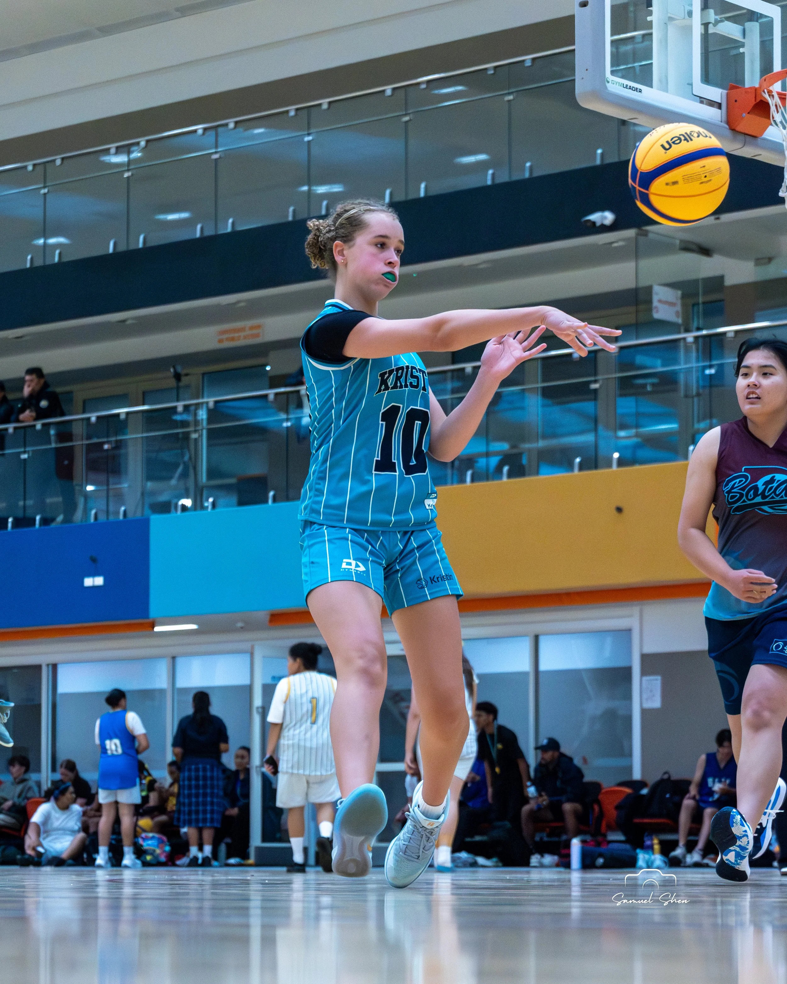 A female volleyball player in a blue and black uniform jumps to hit a volleyball during a match in an indoor gymnasium, with other players and spectators in the background.