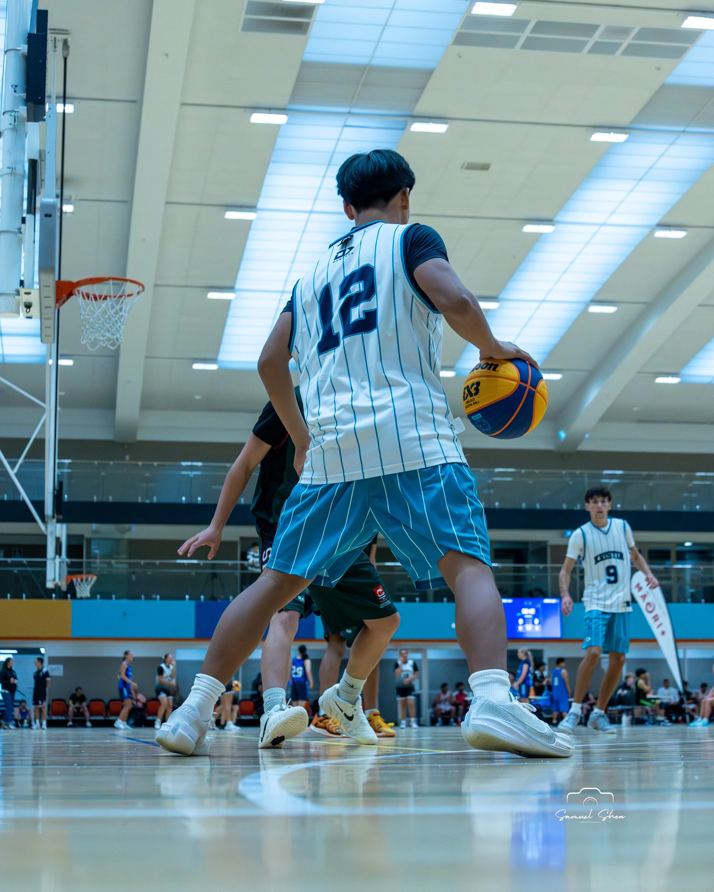 A basketball player in a white numbered jersey with blue pinstripes is in a gym, holding a basketball and preparing to make a move. Other players and spectators are visible in the background.