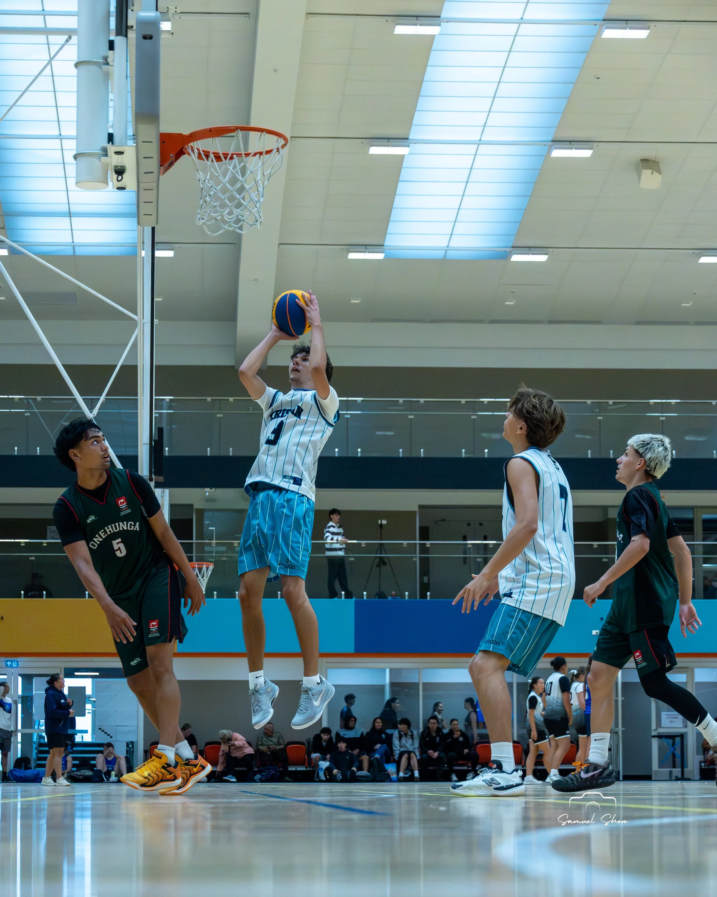 Young male basketball player in mid-air attempting a shot at the hoop, with other players and spectators watching in an indoor gymnasium.