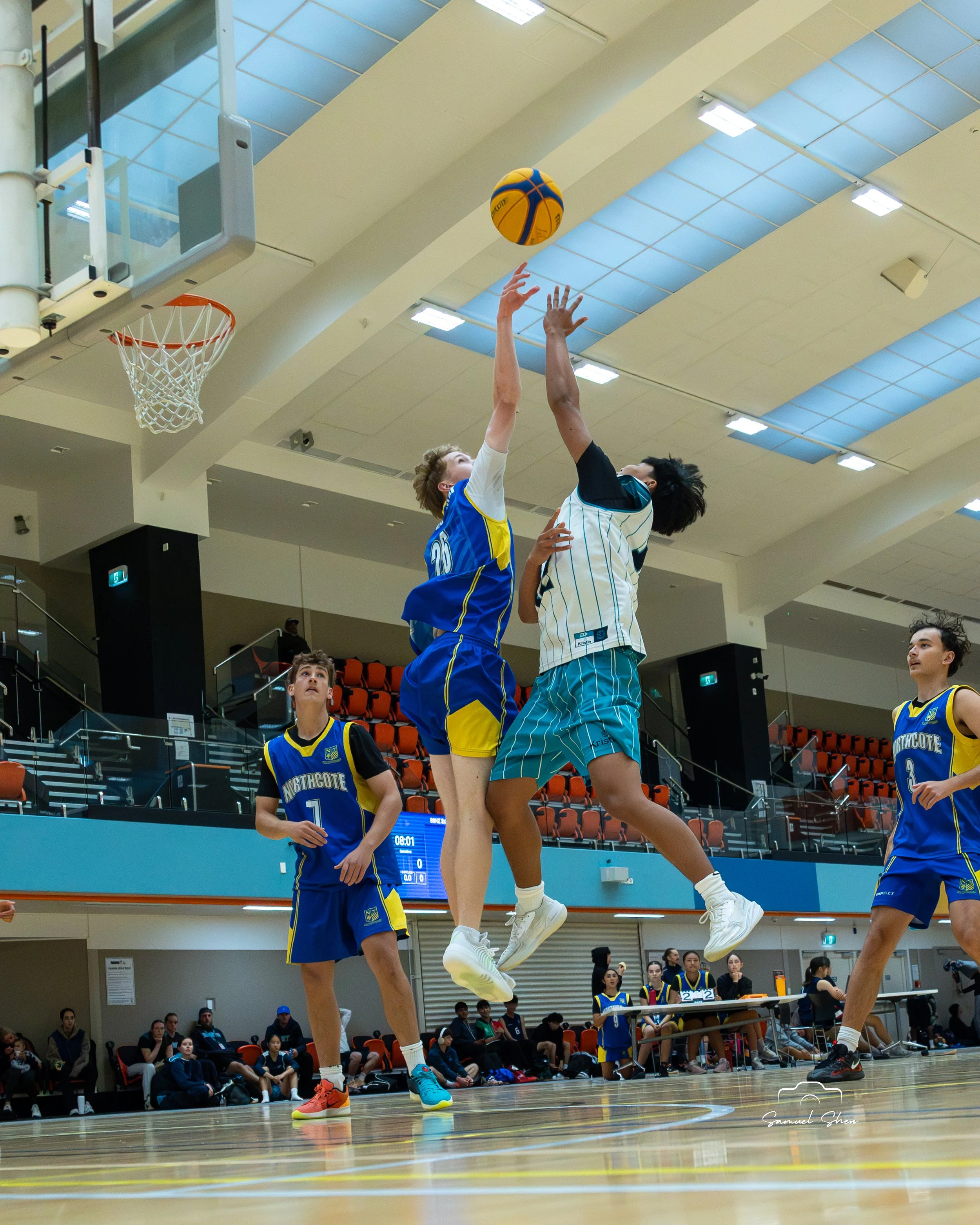Two basketball players compete for the ball mid-air during a game, with one player in a white and teal jersey and the other in a blue and yellow jersey. In the background, spectators sit and stand, watching the game inside a large indoor gymnasium.