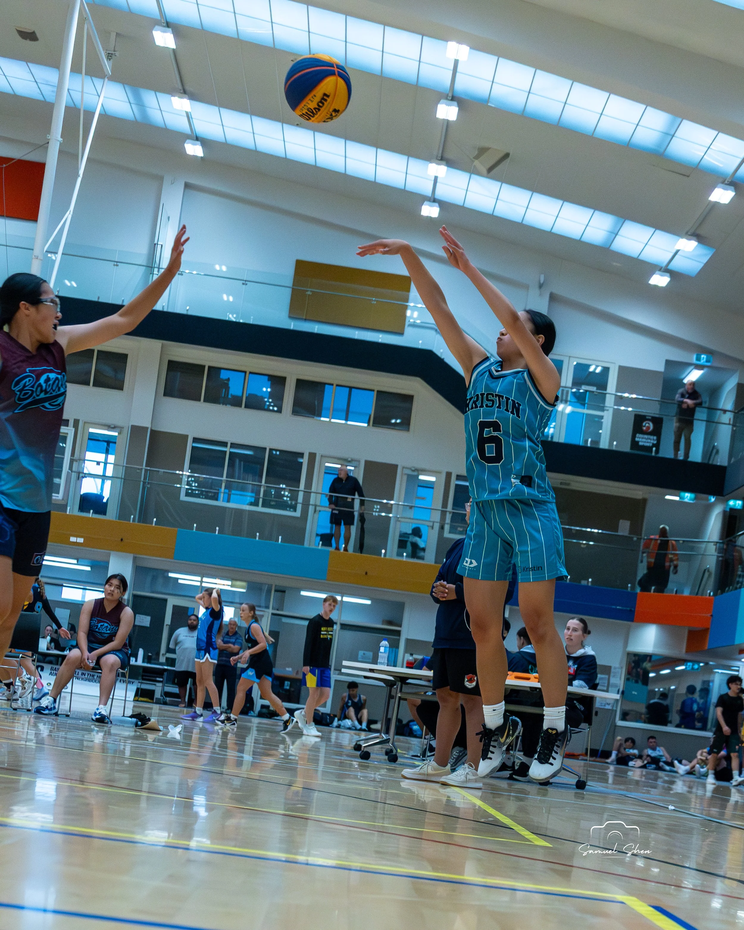 A women playing volleyball in an indoor sports court, with spectators and other players watching in the background.