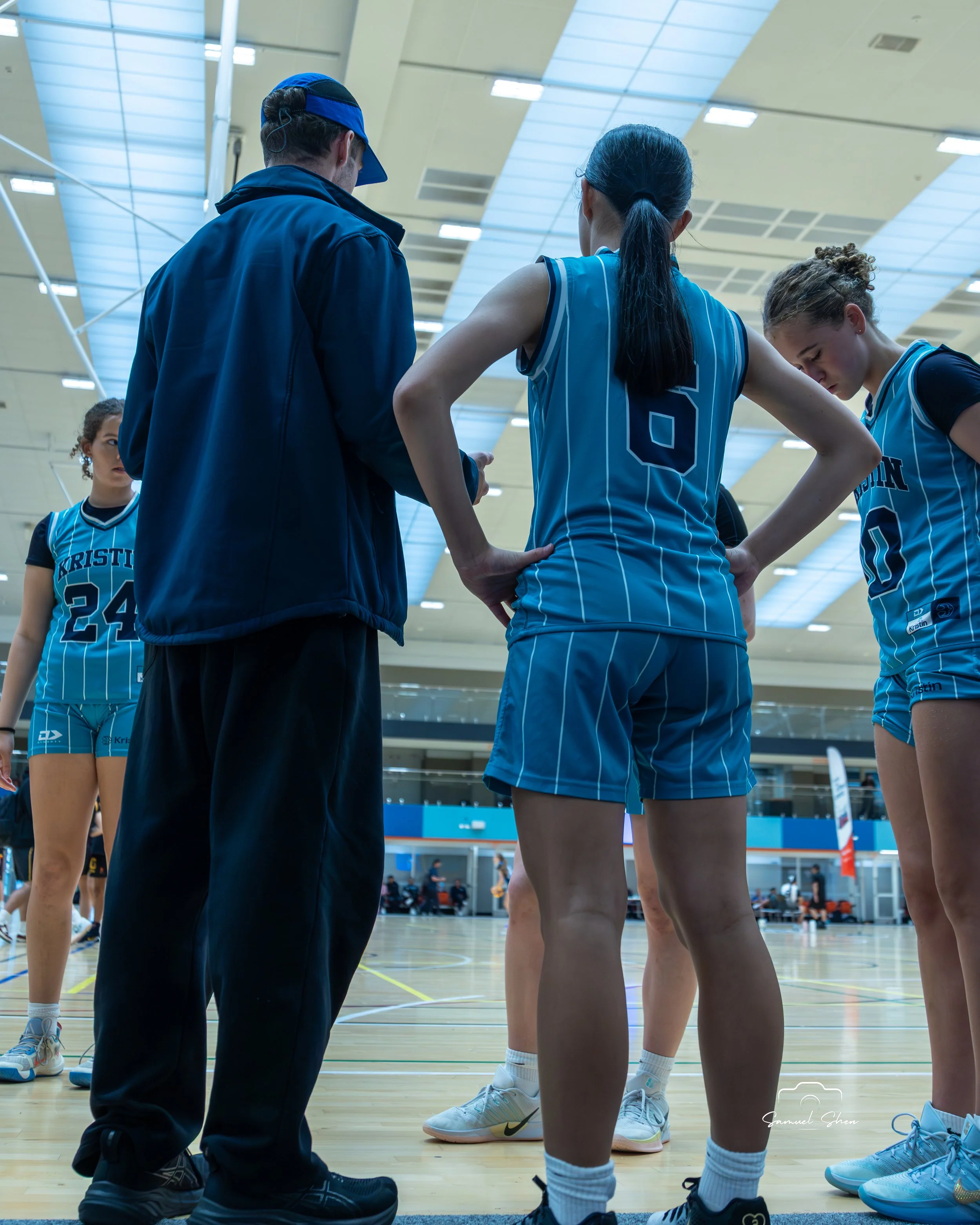 A basketball coach is giving instructions to a female player on the sidelines of an indoor basketball court. Other players are standing nearby, some with heads bowed, wearing blue and black uniforms. The court has bright lighting and a high arched ce