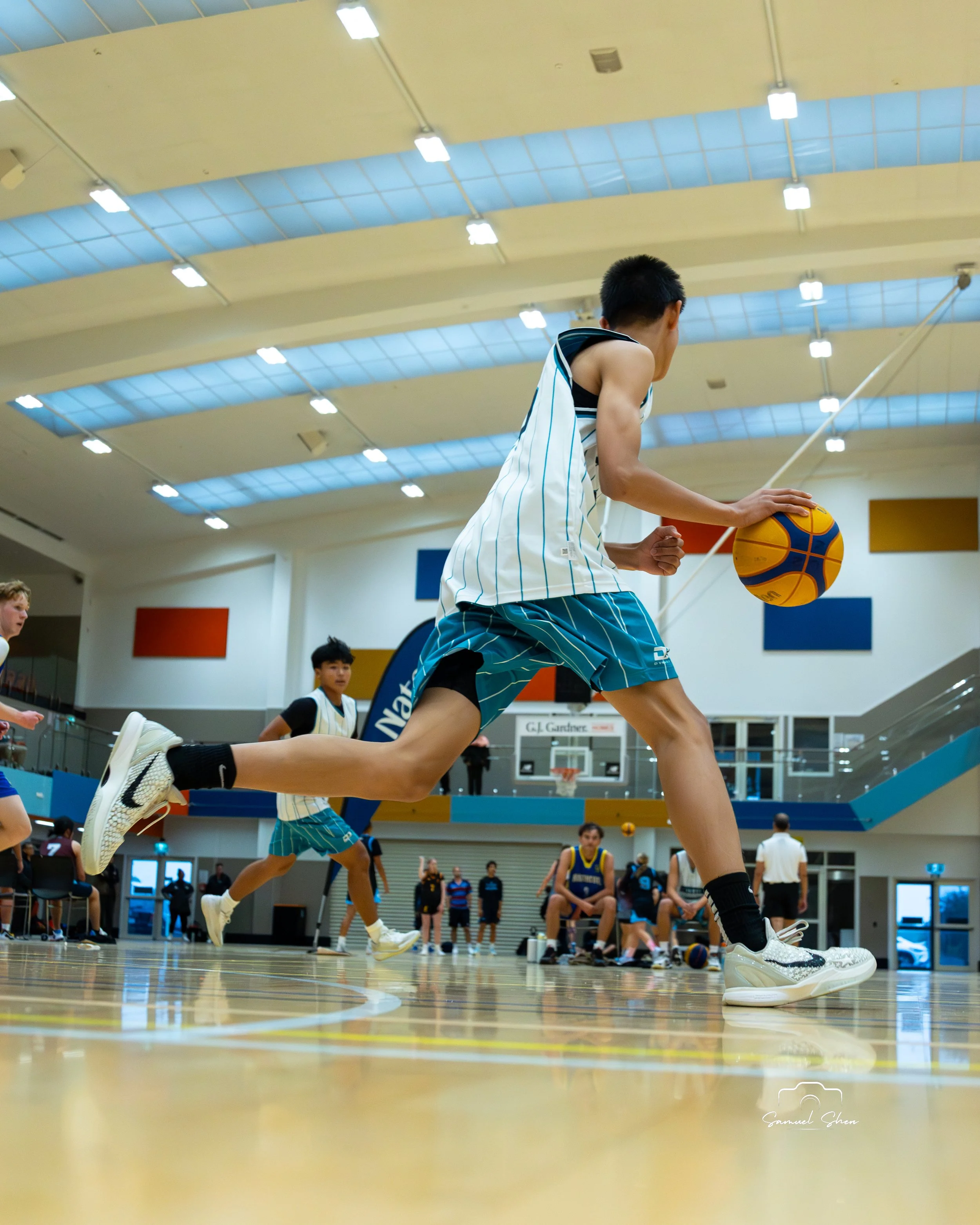 Young male basketball player wearing a striped jersey and shorts dribbling a basketball on an indoor court. Several other players and spectators are visible in the background.