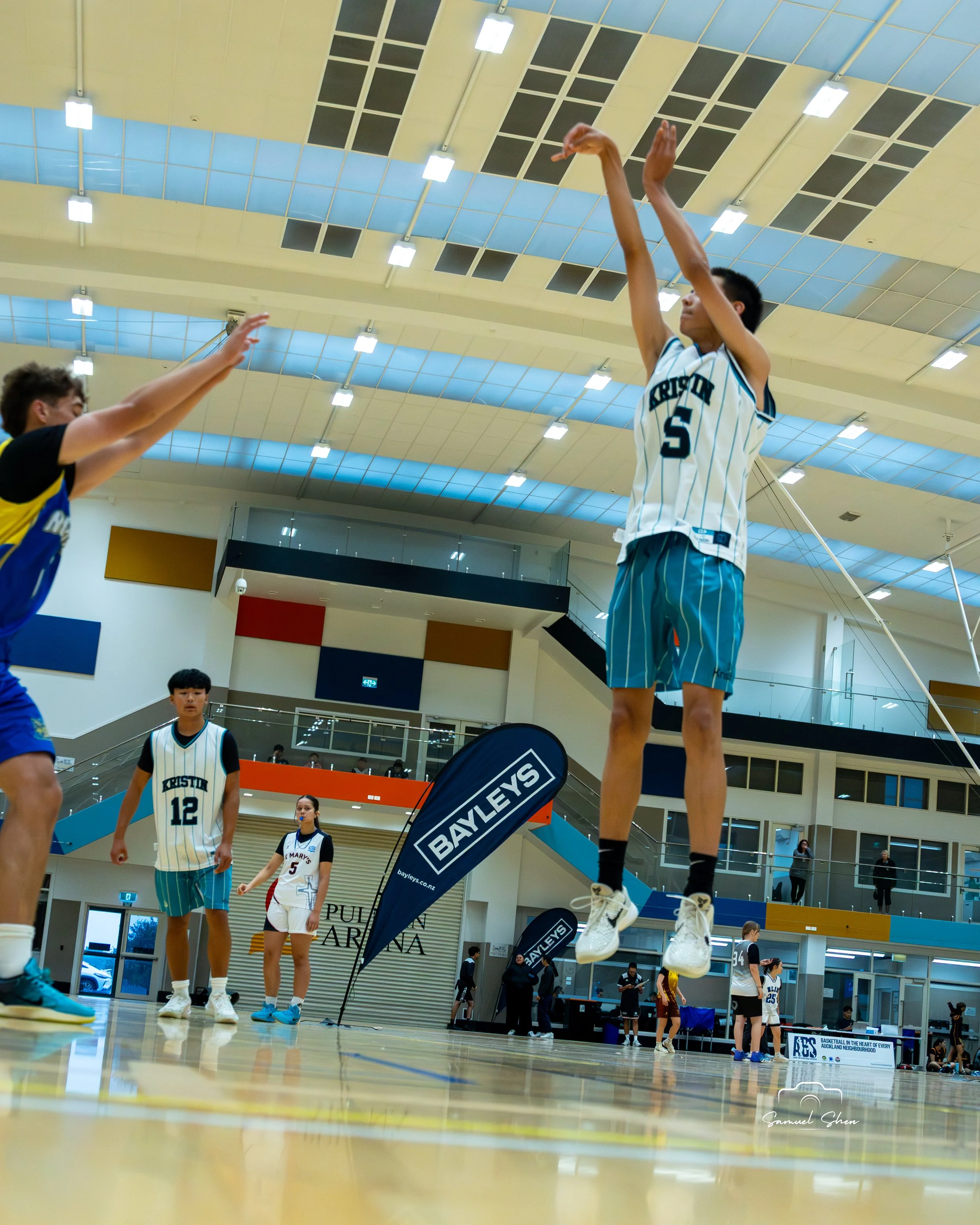 A indoor basketball game in progress with players on the court. Multiple players are visible, some standing and some jumping. The ceiling has large light fixtures and a blue and yellow panel design. There are several banners and signs around the cour