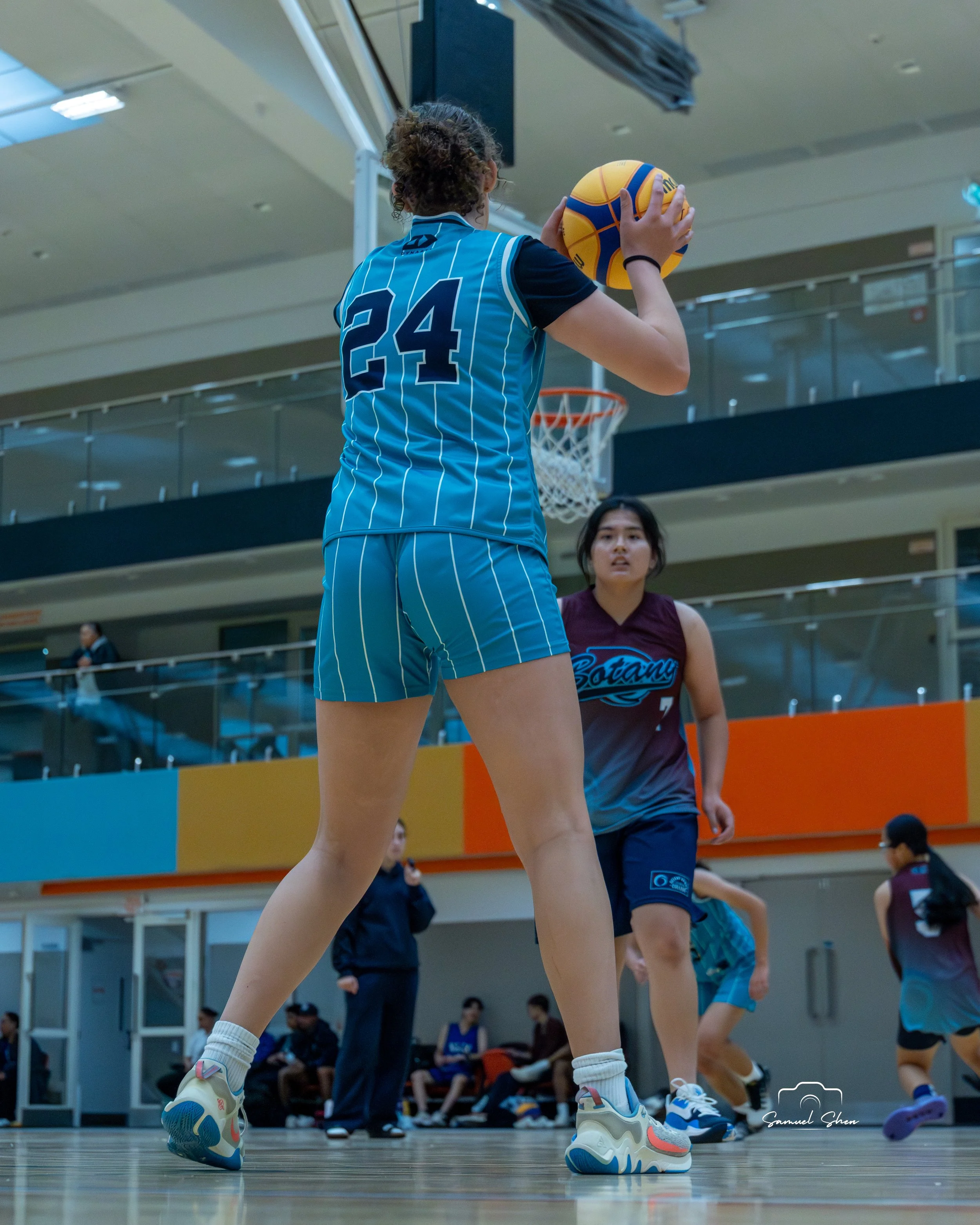 A female basketball player preparing to shoot a ball during a game in an indoor gymnasium, with other players and spectators visible in the background.