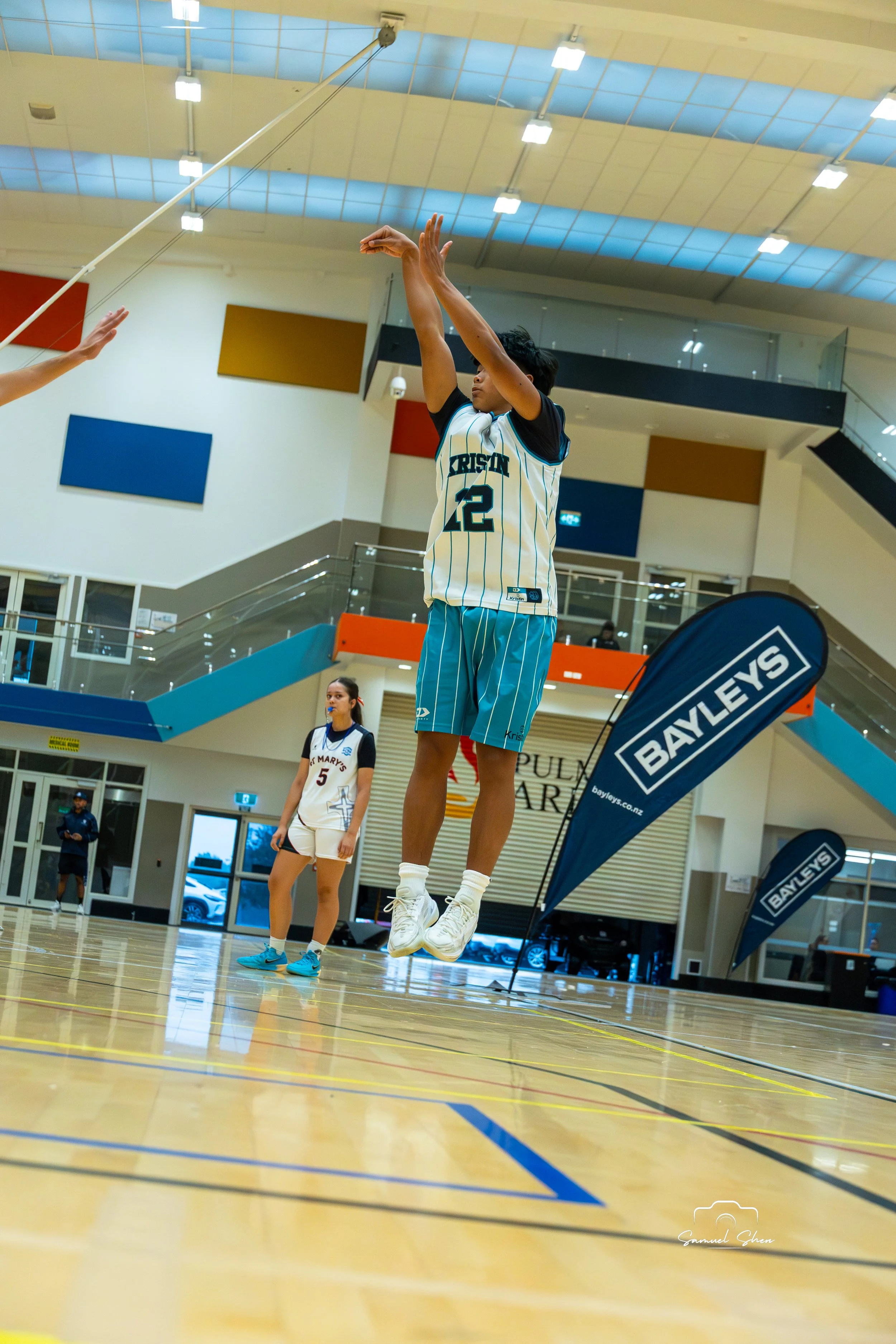 A young woman wearing a turquoise sports uniform with the number 22 jumps in the air during a basketball game at an indoor gym. Two other players, one in a white and black uniform with the number 5, and a coach or referee, are visible in the backgrou