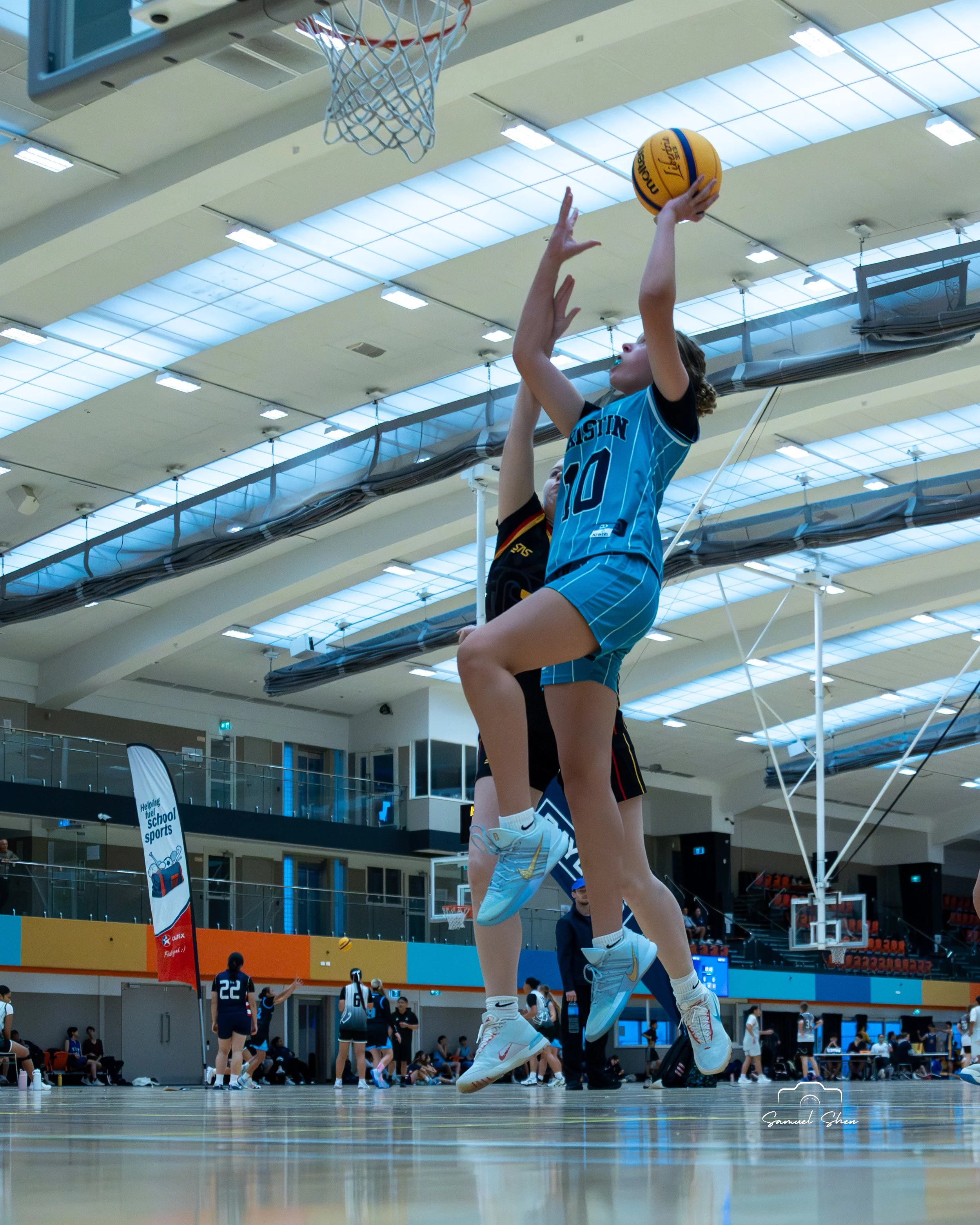 A young female basketball player in a blue jersey with the number 10 jumping to take a shot during a game in an indoor gymnasium, with other players and spectators visible in the background.