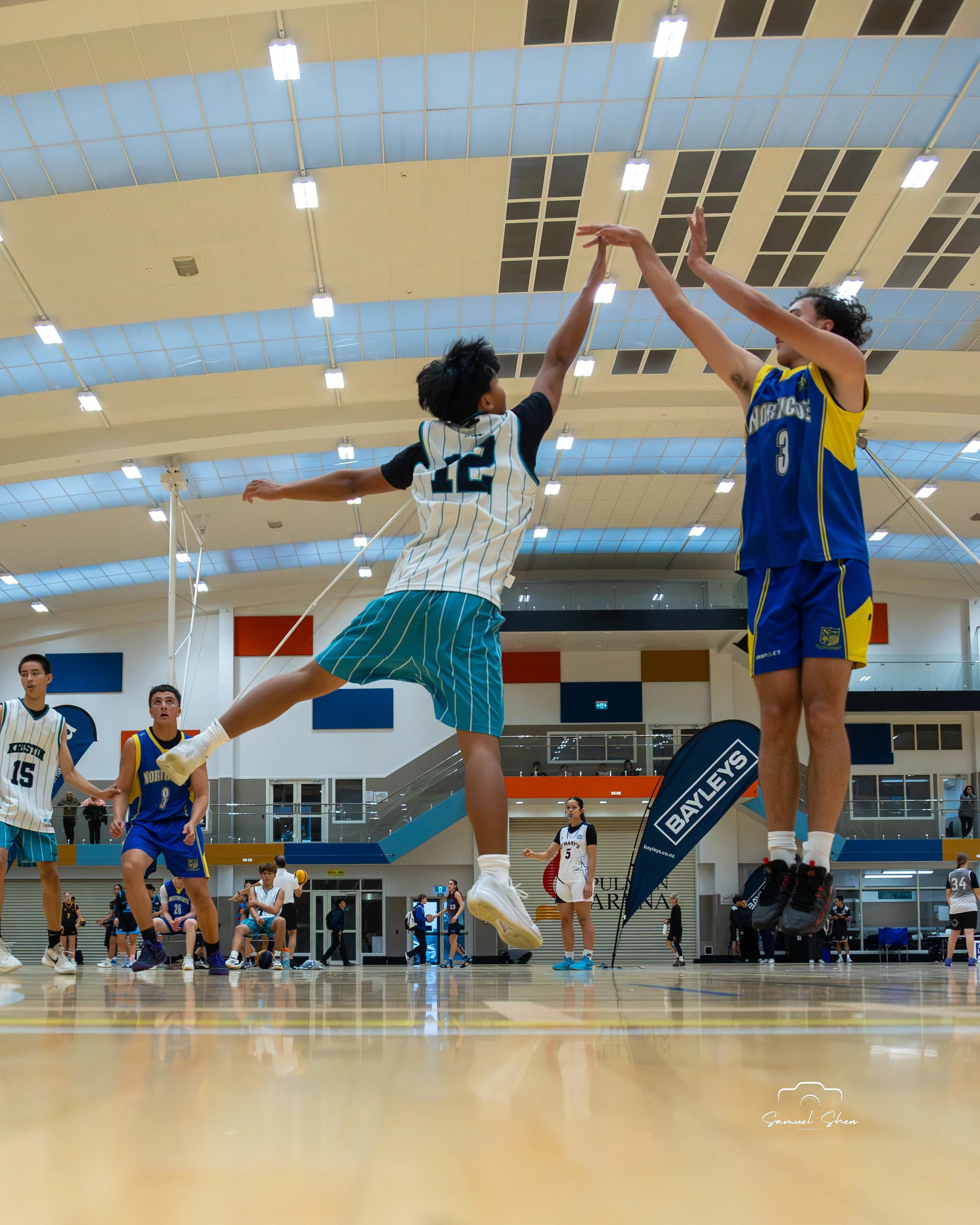 Indoor volleyball game with players in mid-action. Two players are jumping to reach the ball, with one in a white and black uniform and the other in a blue and yellow uniform. Multiple players are visible on the court, some preparing to play, others 