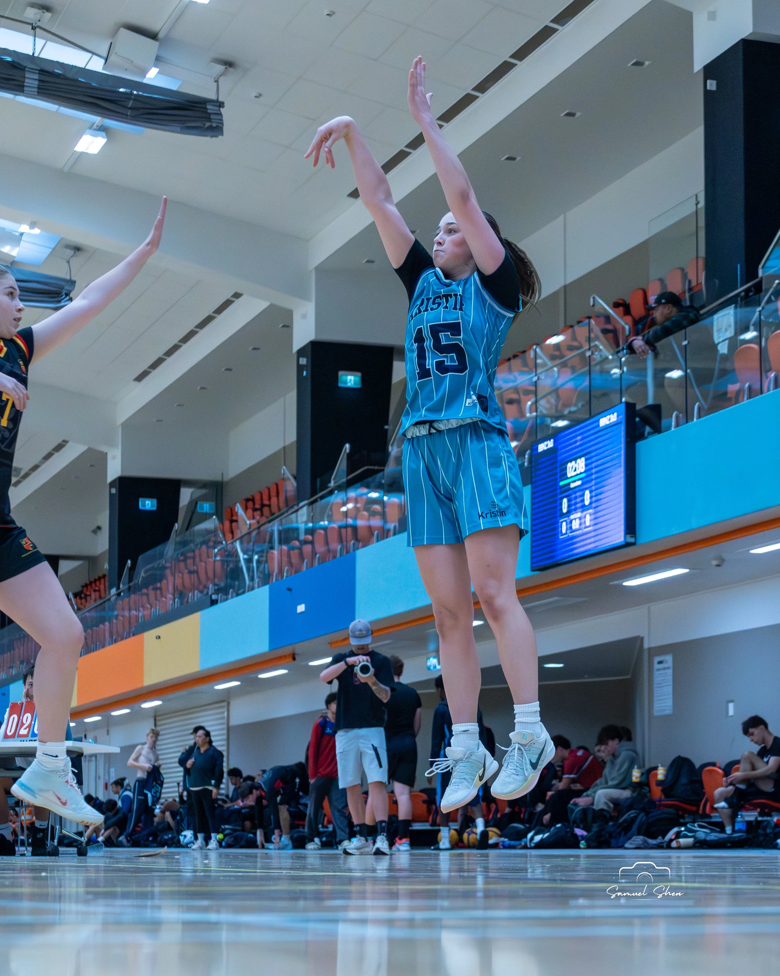 Female volleyball player wearing a blue jersey with the number 15 jumps for a serve during a game in an indoor stadium, with spectators and other players in the background.