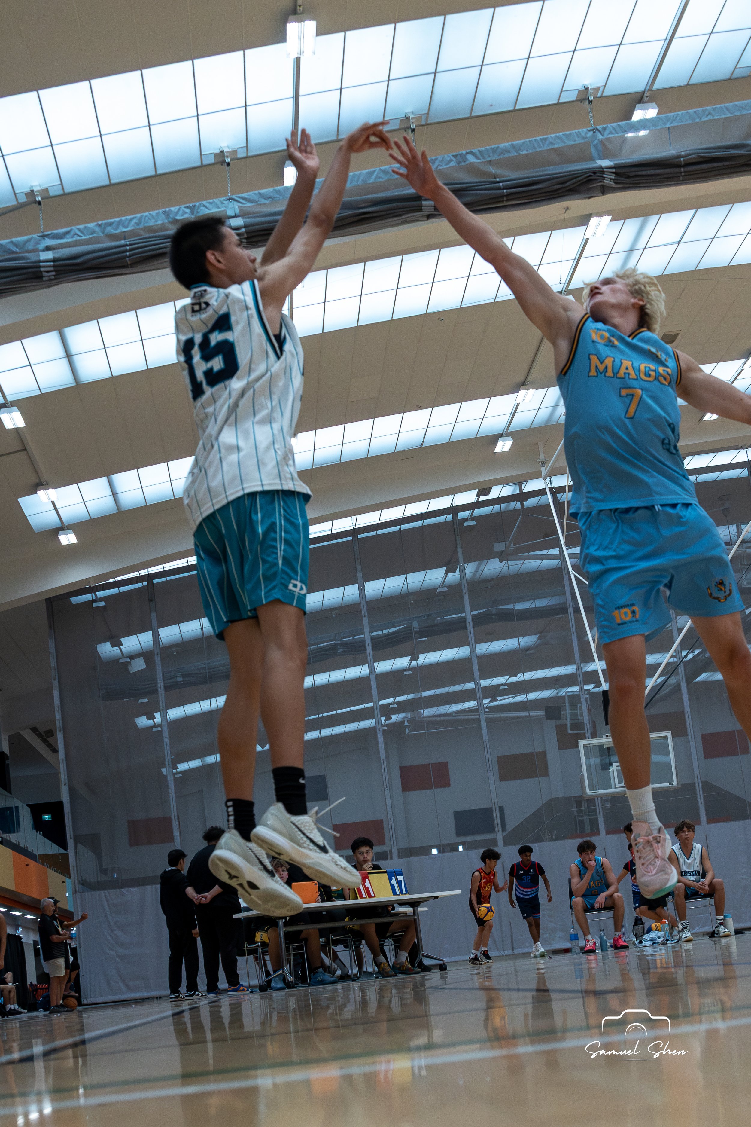 Two basketball players are jumping for the ball in a game, with players and officials on the court and spectators in the background inside a bright gymnasium.