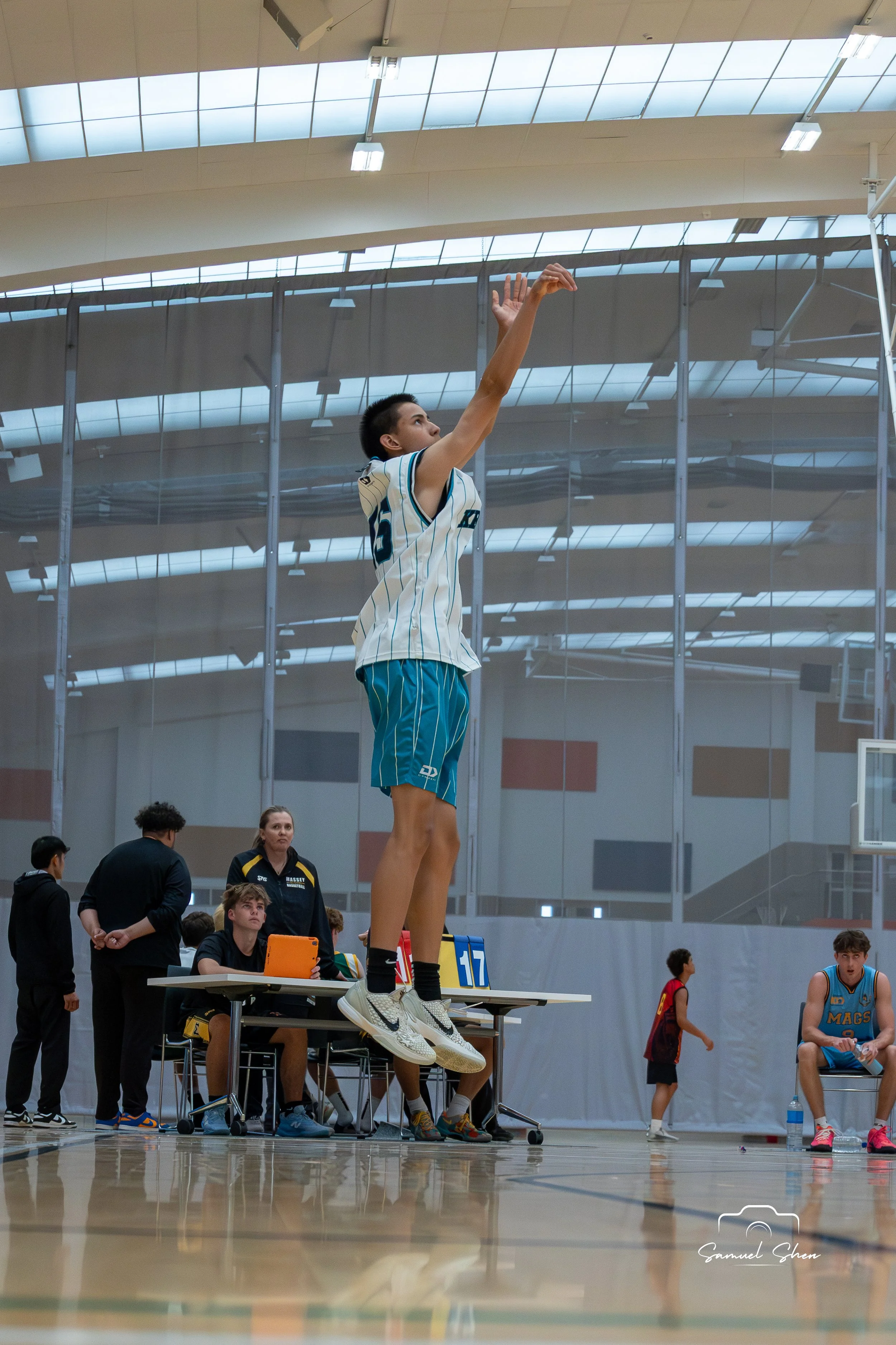 An athlete in a striped jersey jumps to take a shot during a basketball game in an indoor gym, with spectators and officials watching.