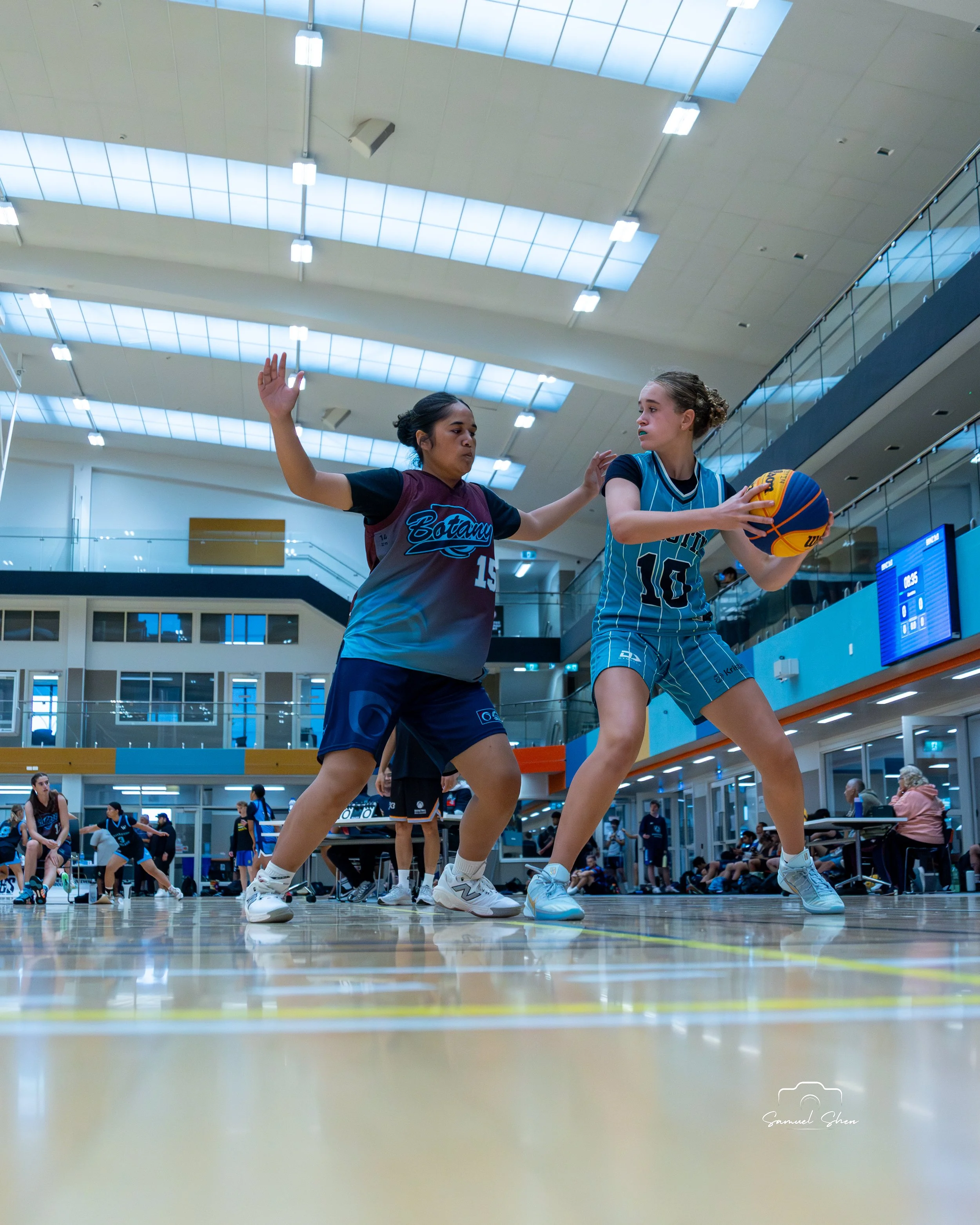 Two women playing basketball indoors, with others sitting on benches and watching in the background.